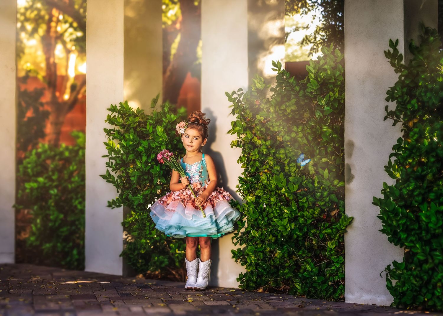 A little girl in a colorful tiered dress and white boots stands between lush green bushes in golden light.