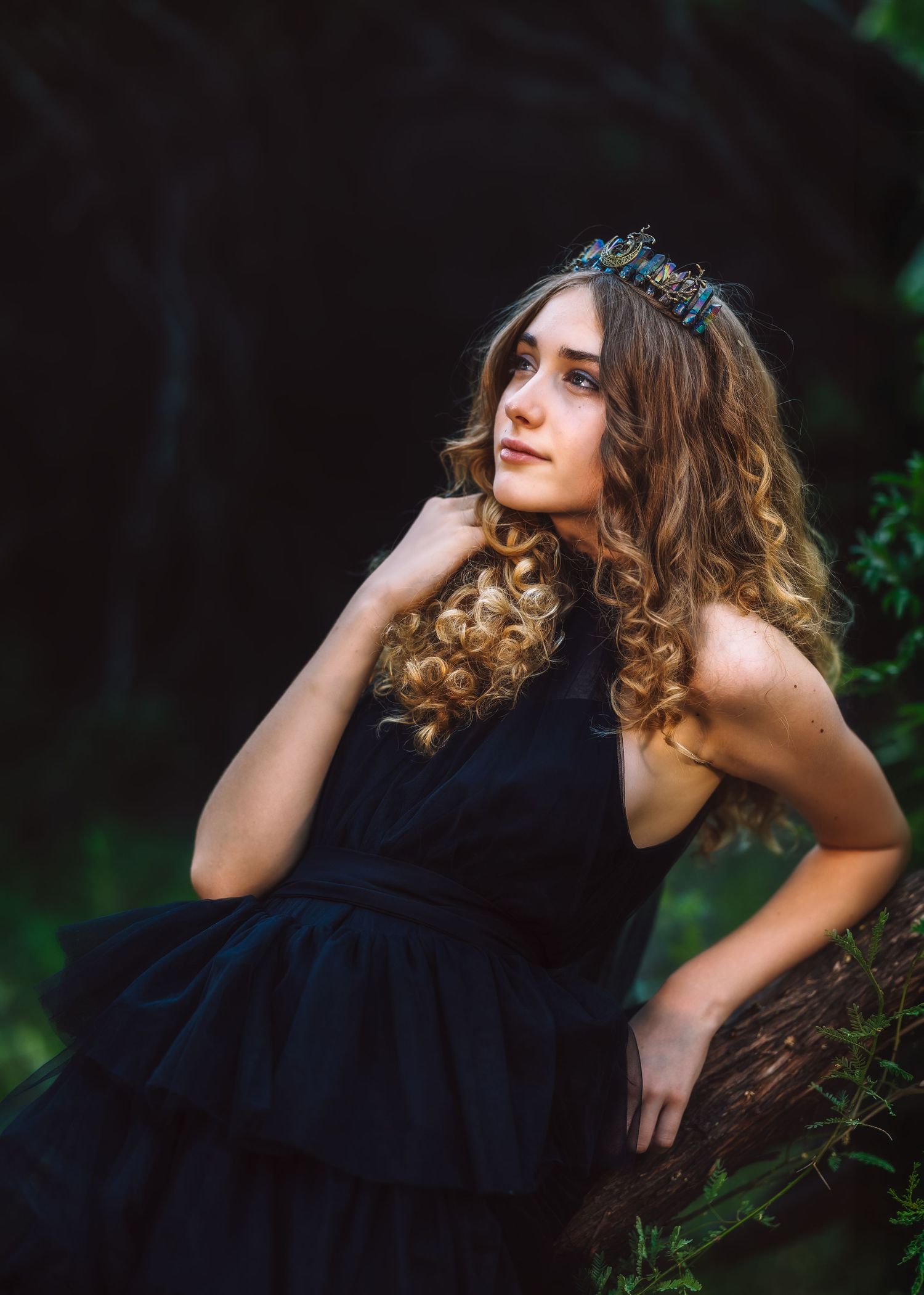 Young woman in black dress and crown poses gracefully in a dark forest setting with curly hair.