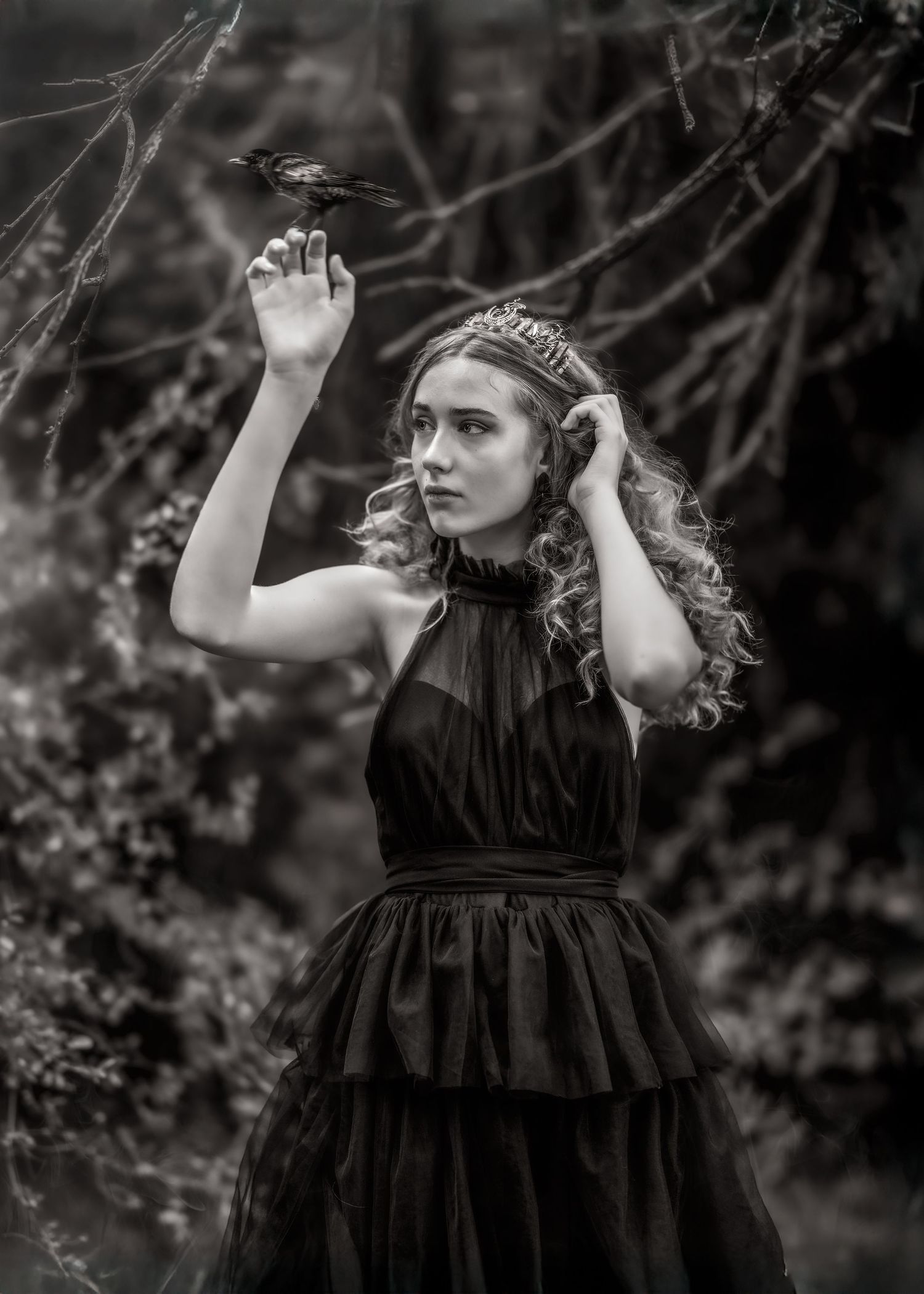 Black and white photo of a young woman in a black dress holding a bird on her hand in a dark forest setting.