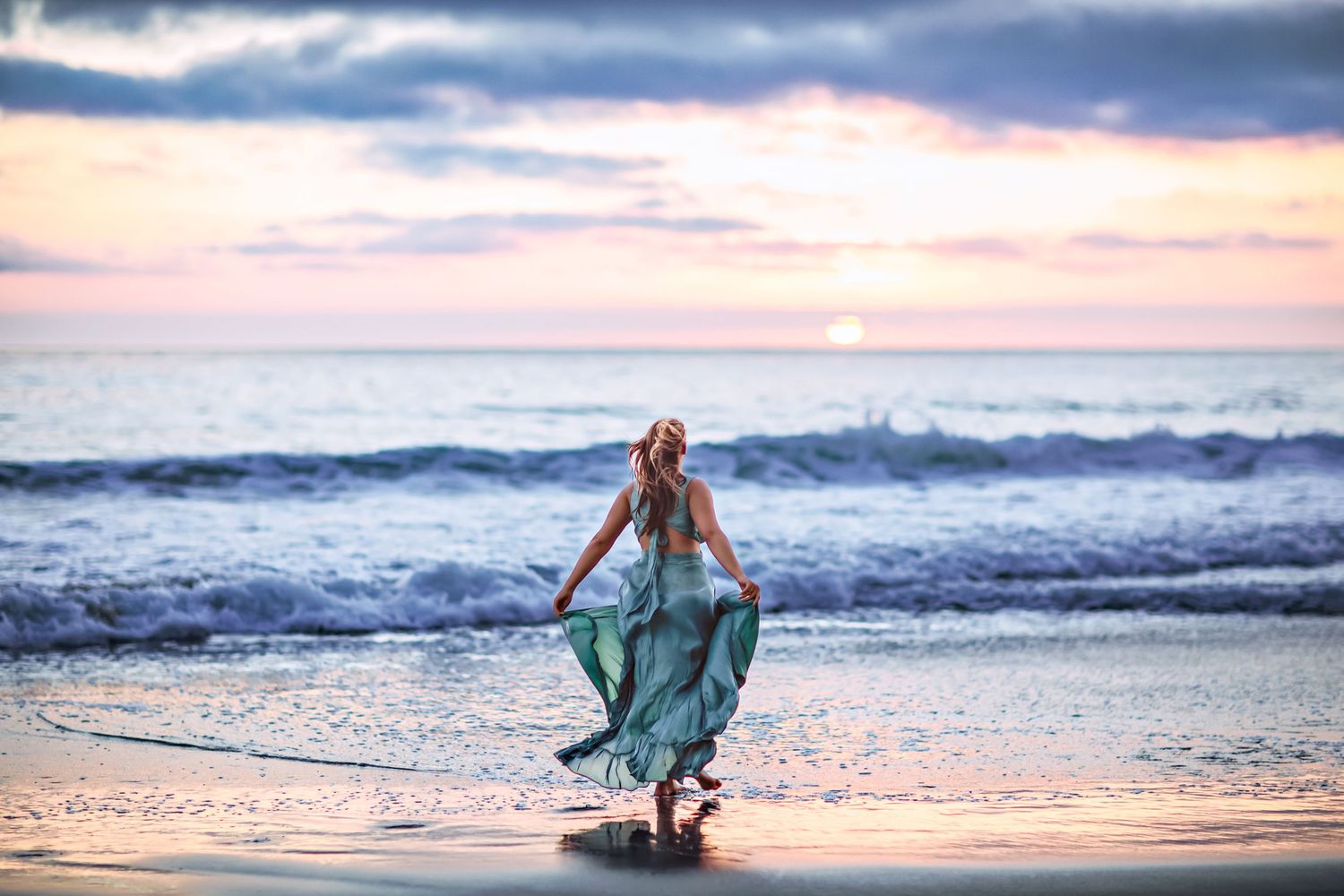 Woman in teal dress walking toward ocean waves at sunset.
