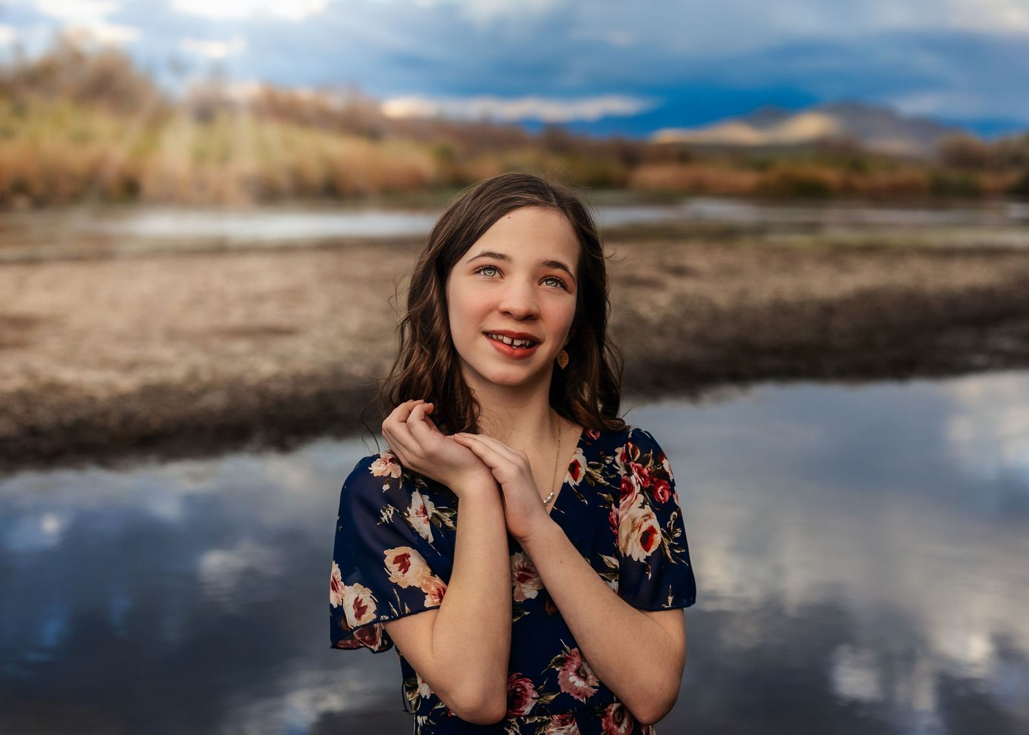 A smiling young girl in a floral dress poses with hands clasped near a reflective lake under dramatic cloudy skies.