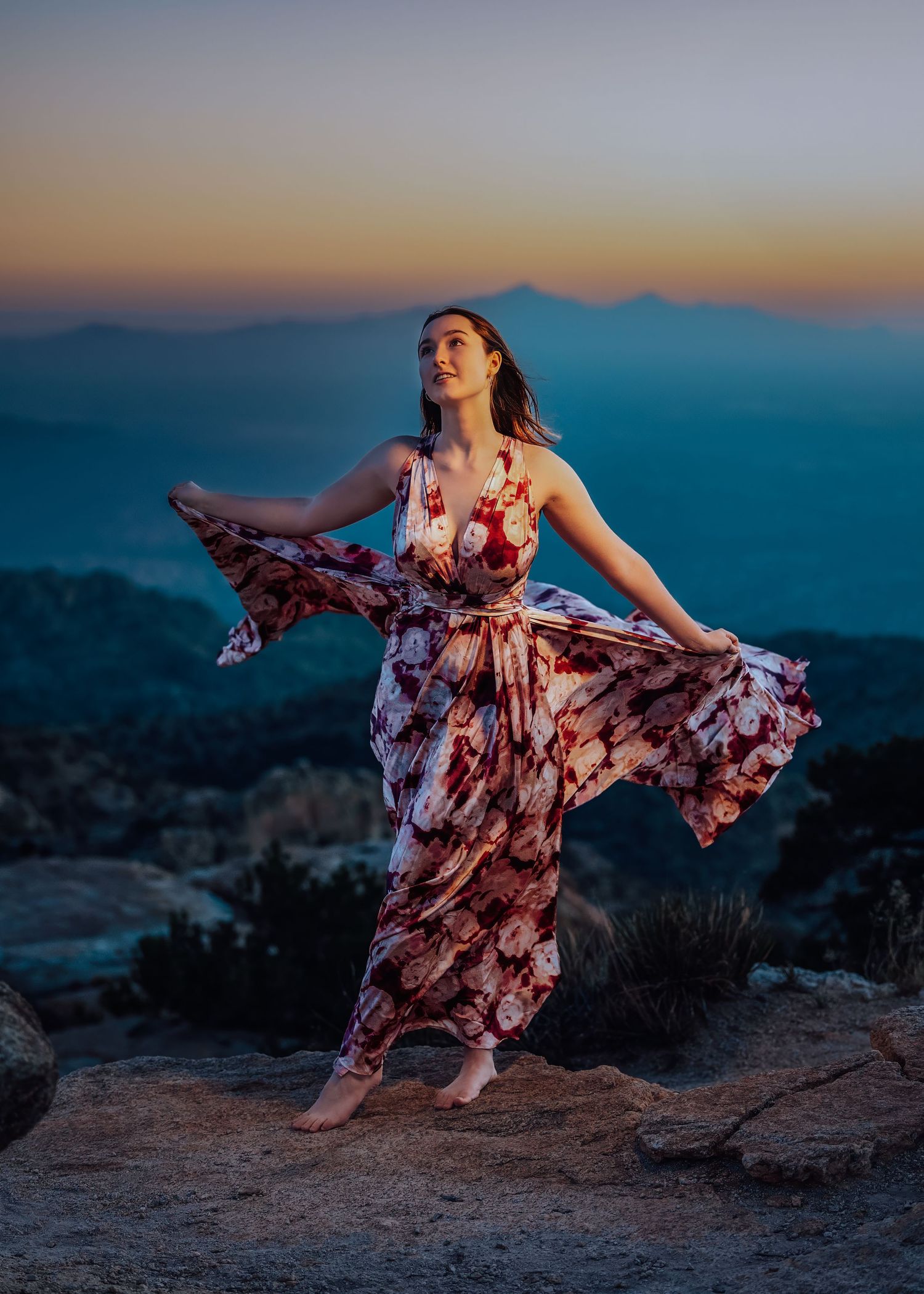 Barefoot woman in flowing floral maxi dress posing on rocks at blue hour mountain sunset.