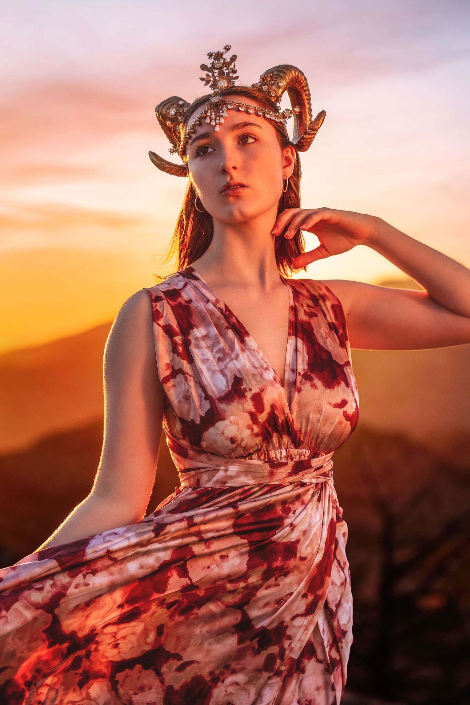 Woman in floral dress wearing decorative ram horn headpiece poses at vibrant mountain sunset.
