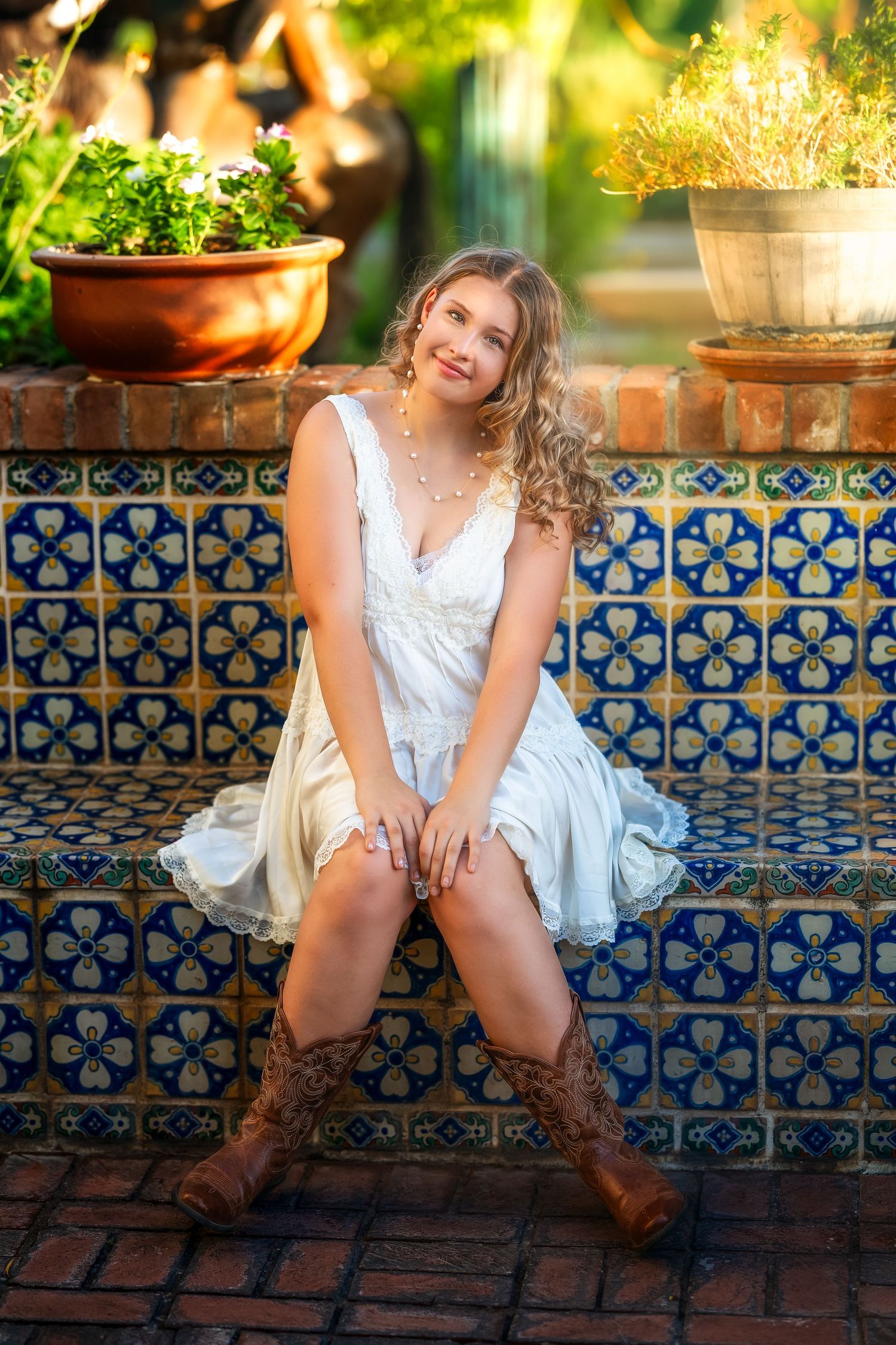 Young woman in white dress and cowboy boots sitting on colorful tiled bench surrounded by potted plants.
