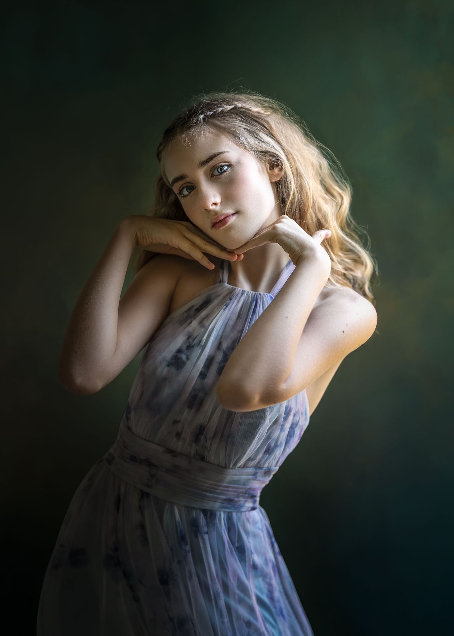 Young woman in a floral blue dress poses gracefully with hands near chin against a dark green background.