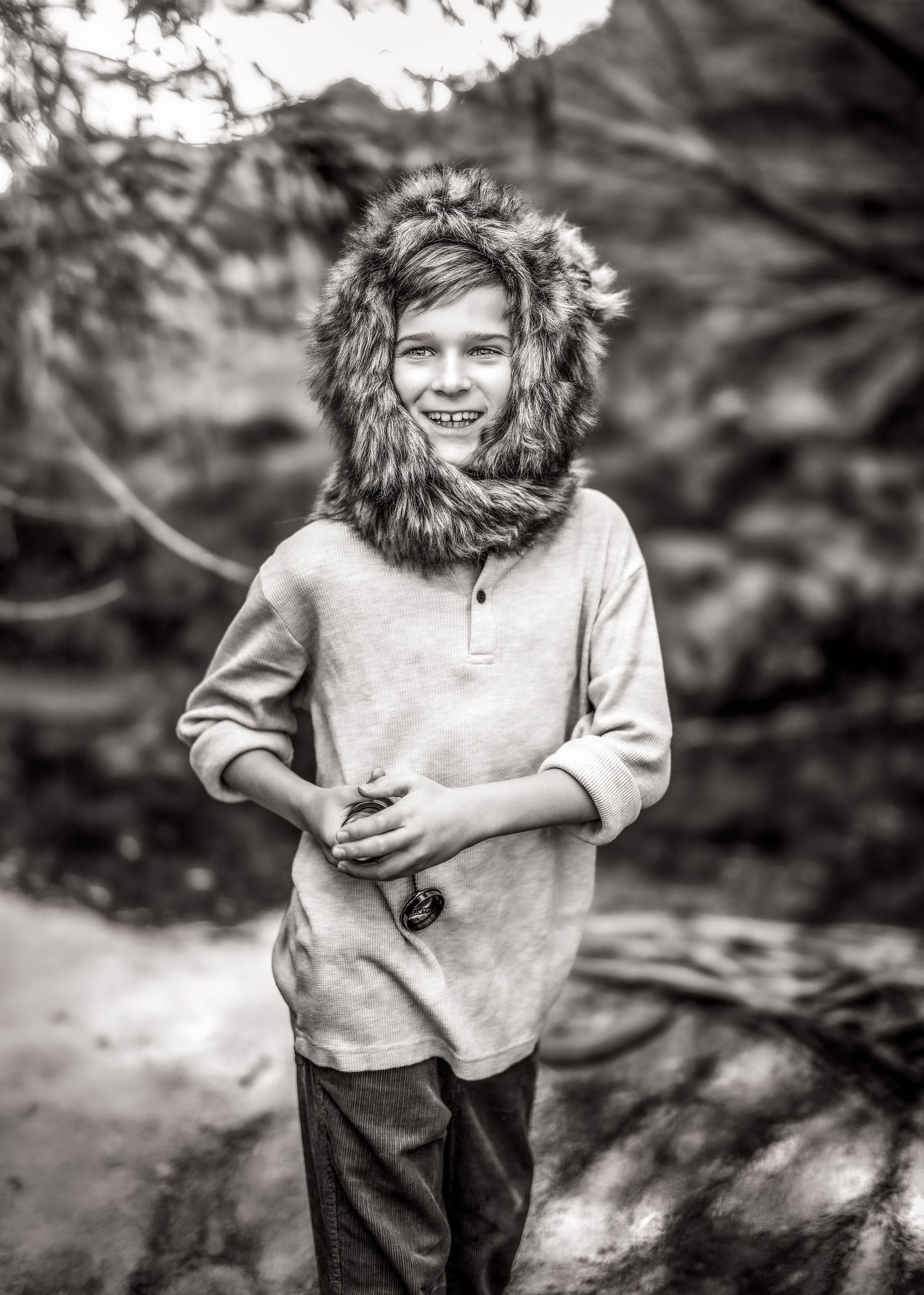 A smiling boy in a furry hooded hat poses outdoors in a black and white portrait with trees behind him.