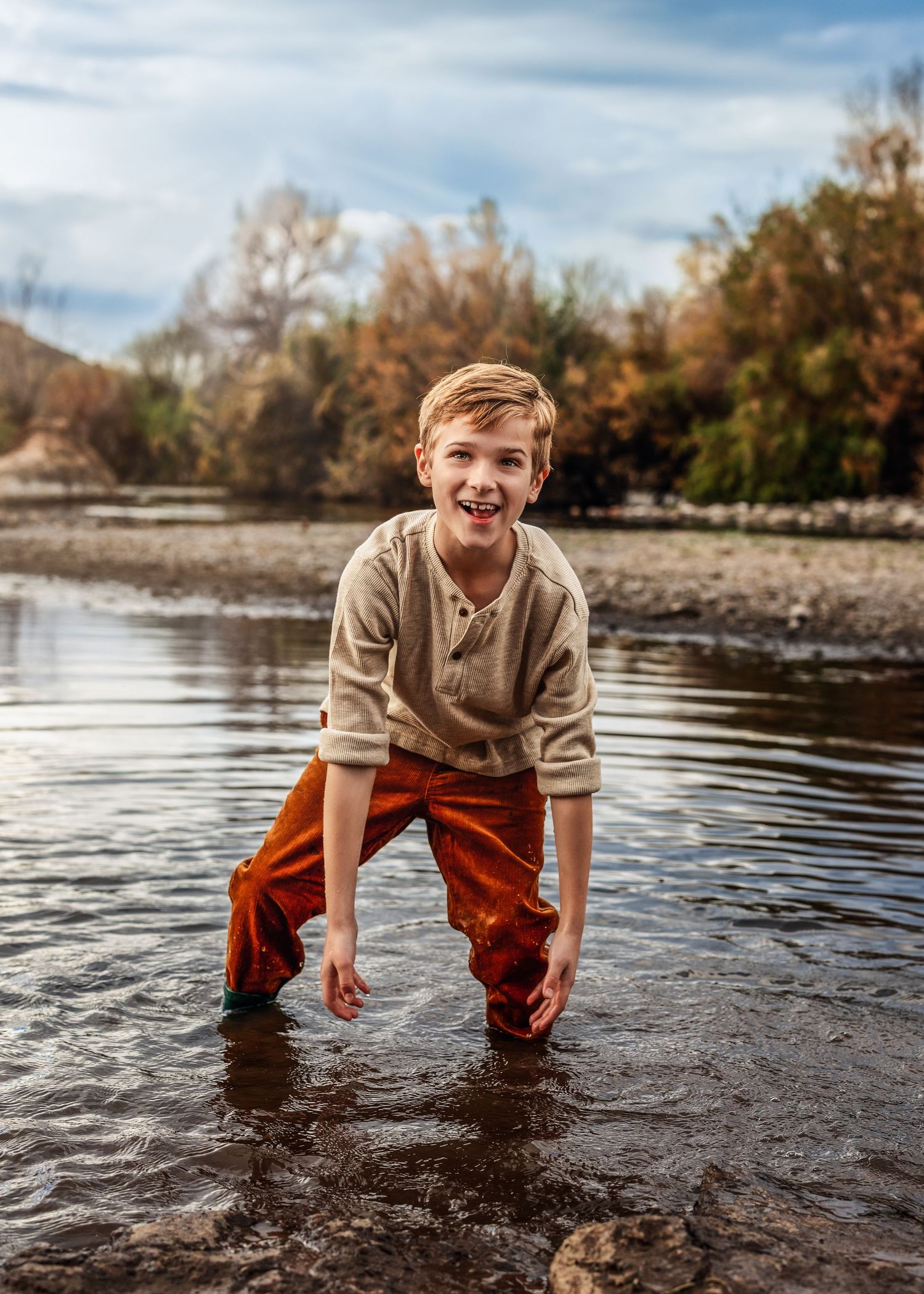 Laughing boy in rust orange pants splashes and plays in a shallow river surrounded by autumn trees.