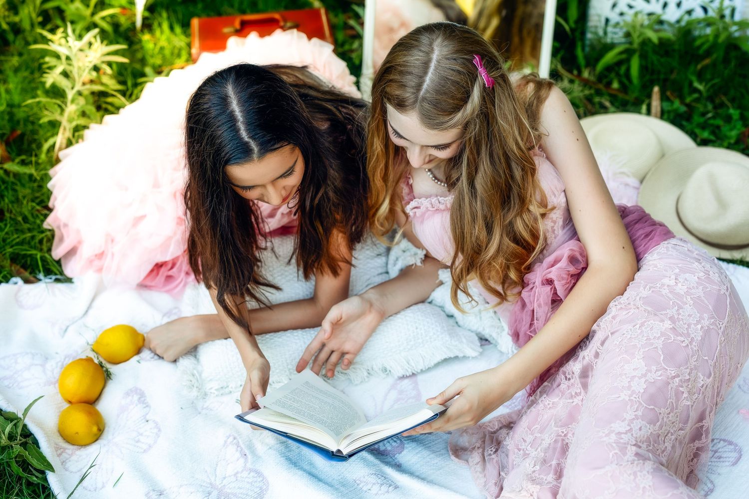 Two girls in pretty dresses reading a book together on a blanket outdoors with lemons nearby.