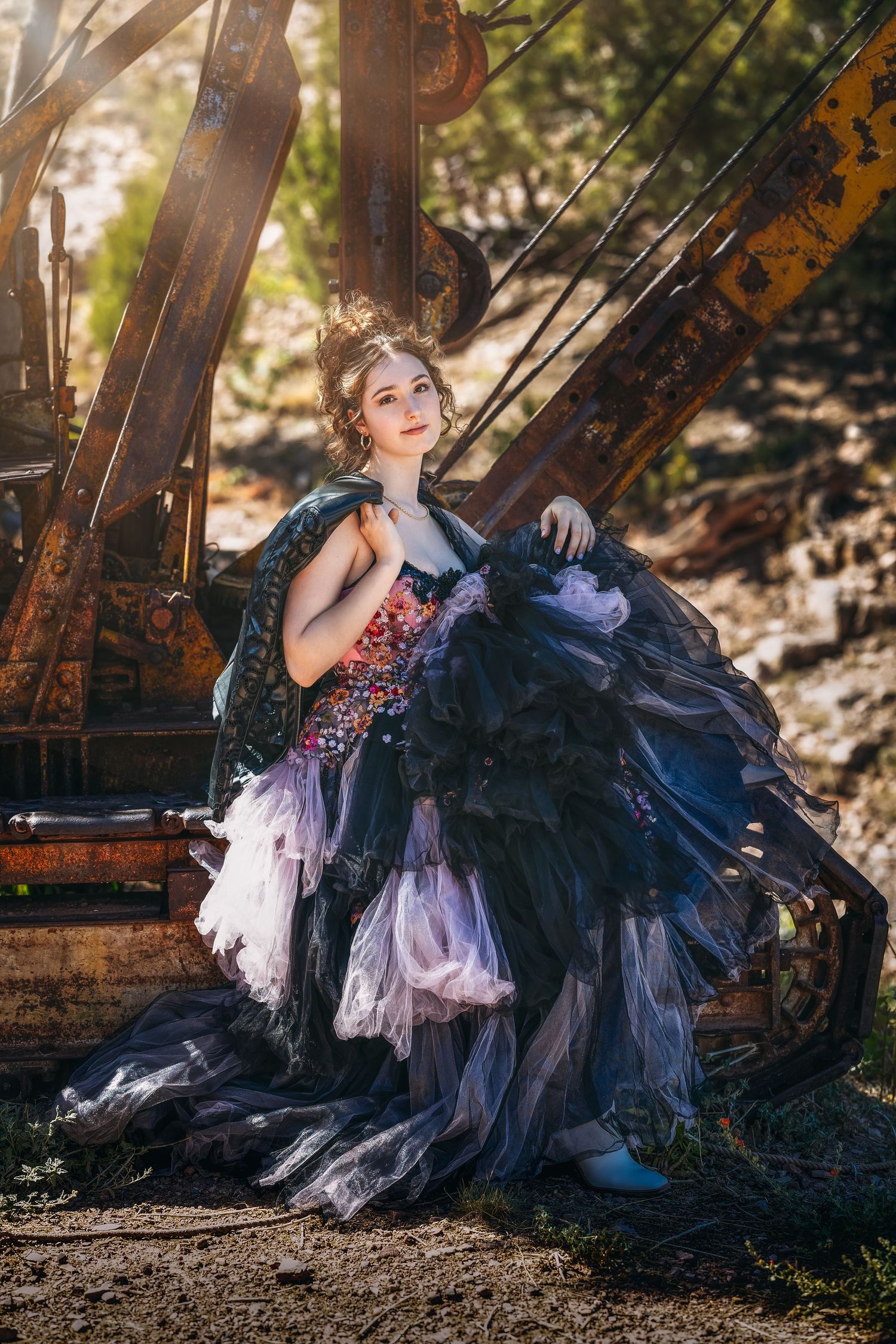 Young woman in an elaborate black and pink ballgown poses beside rusty industrial machinery in golden sunlight.