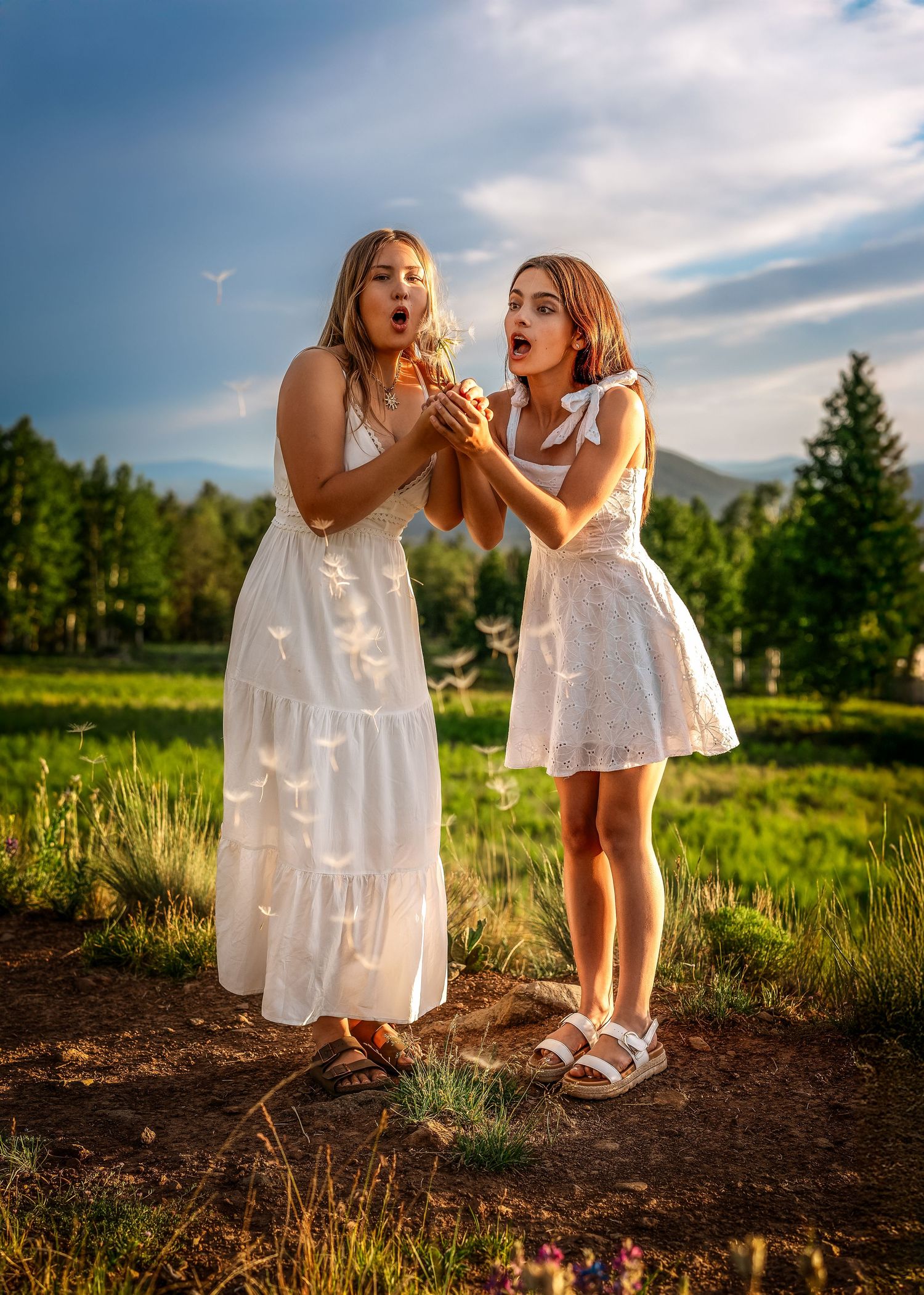 Two young women in white dresses blowing dandelion seeds in a scenic green meadow.