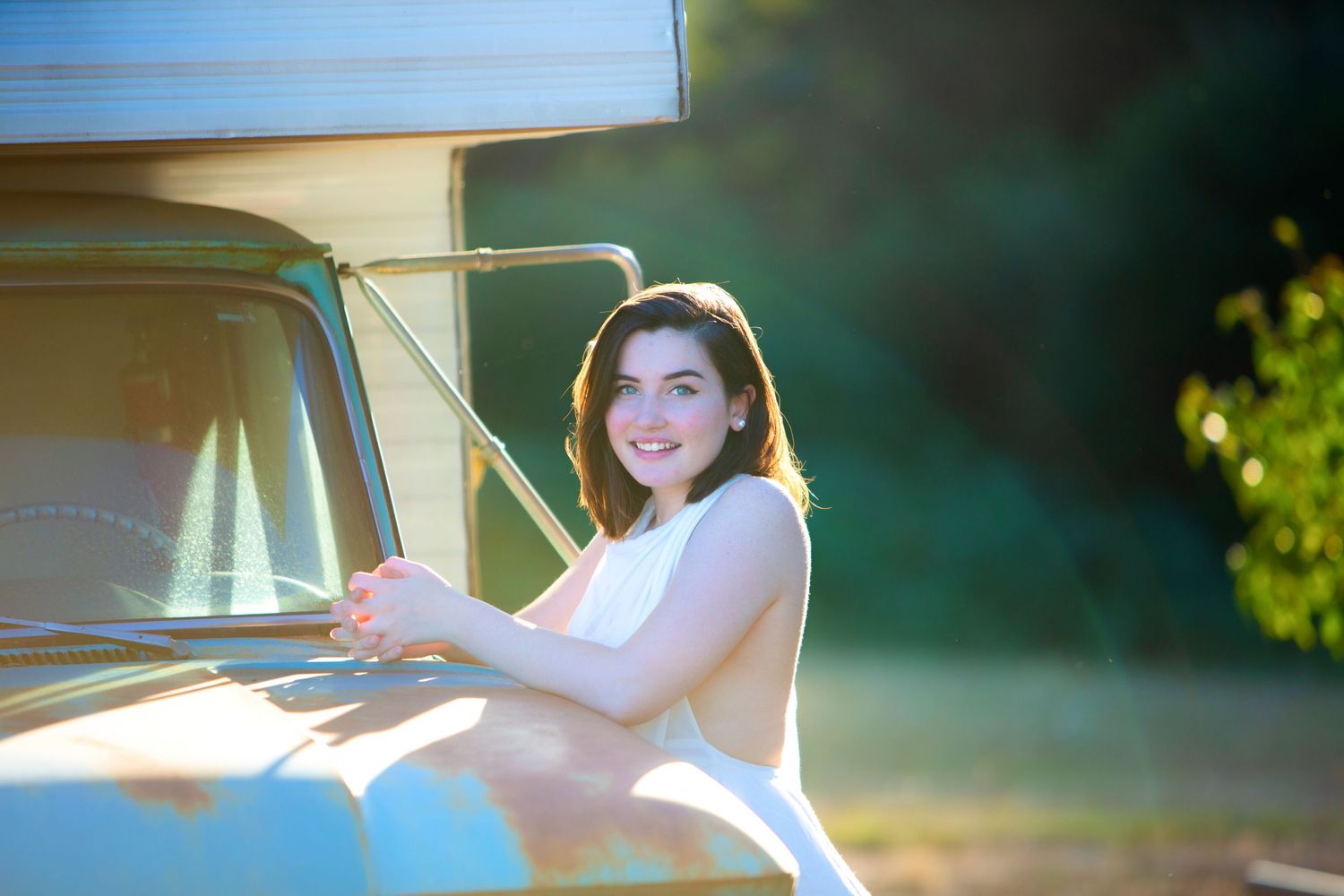 Smiling young woman in white dress posing beside classic blue pickup truck in sunlight.