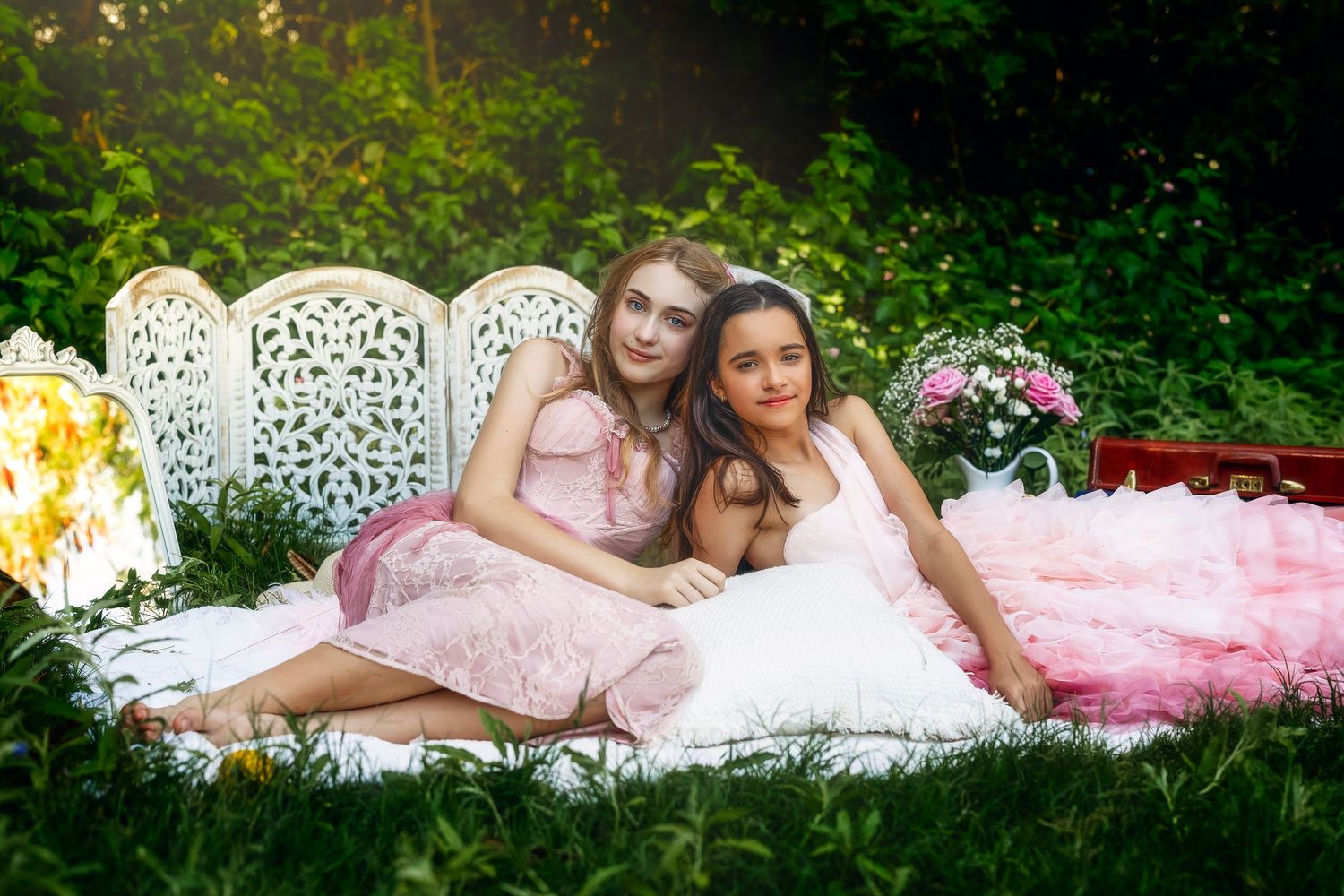 Two young women in pink dresses pose together on a white ornate bench surrounded by lush green garden foliage.