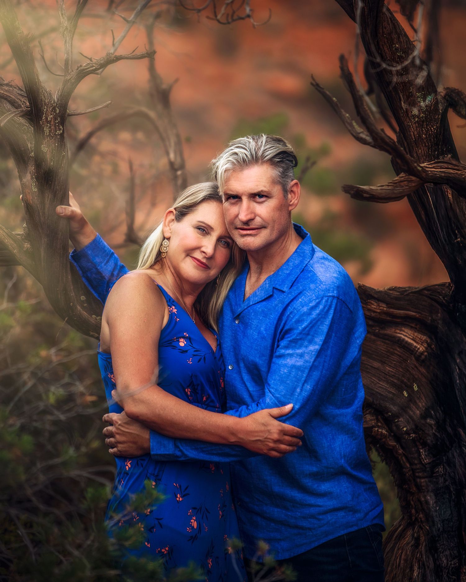 Couple in matching blue outfits embracing near a twisted tree in a warm desert landscape.