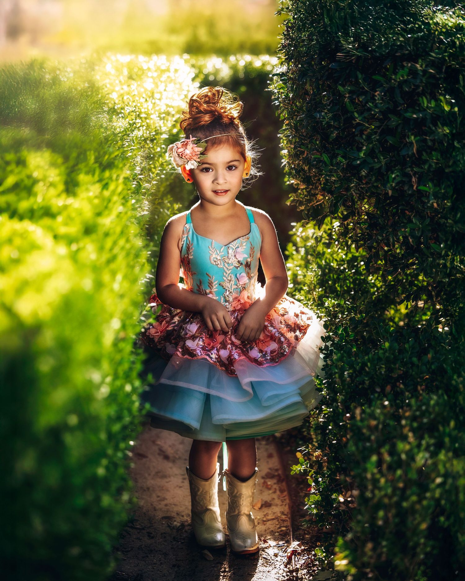 Young girl in teal and pink ruffled dress with cowboy boots walking through lush green garden hedges.