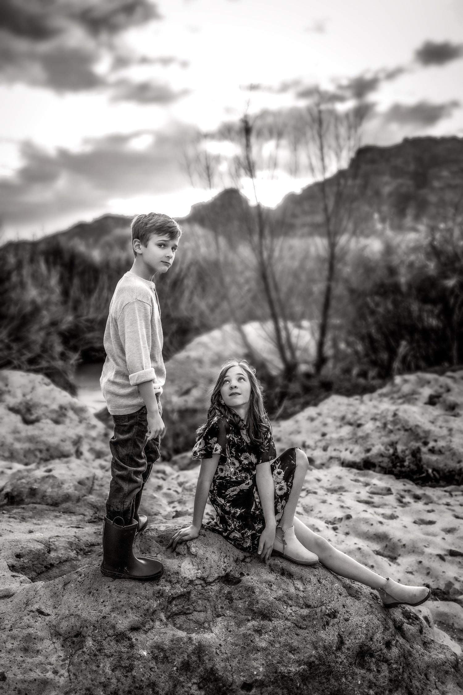Black and white photo of two children posing on rocks with dramatic mountain landscape behind them.