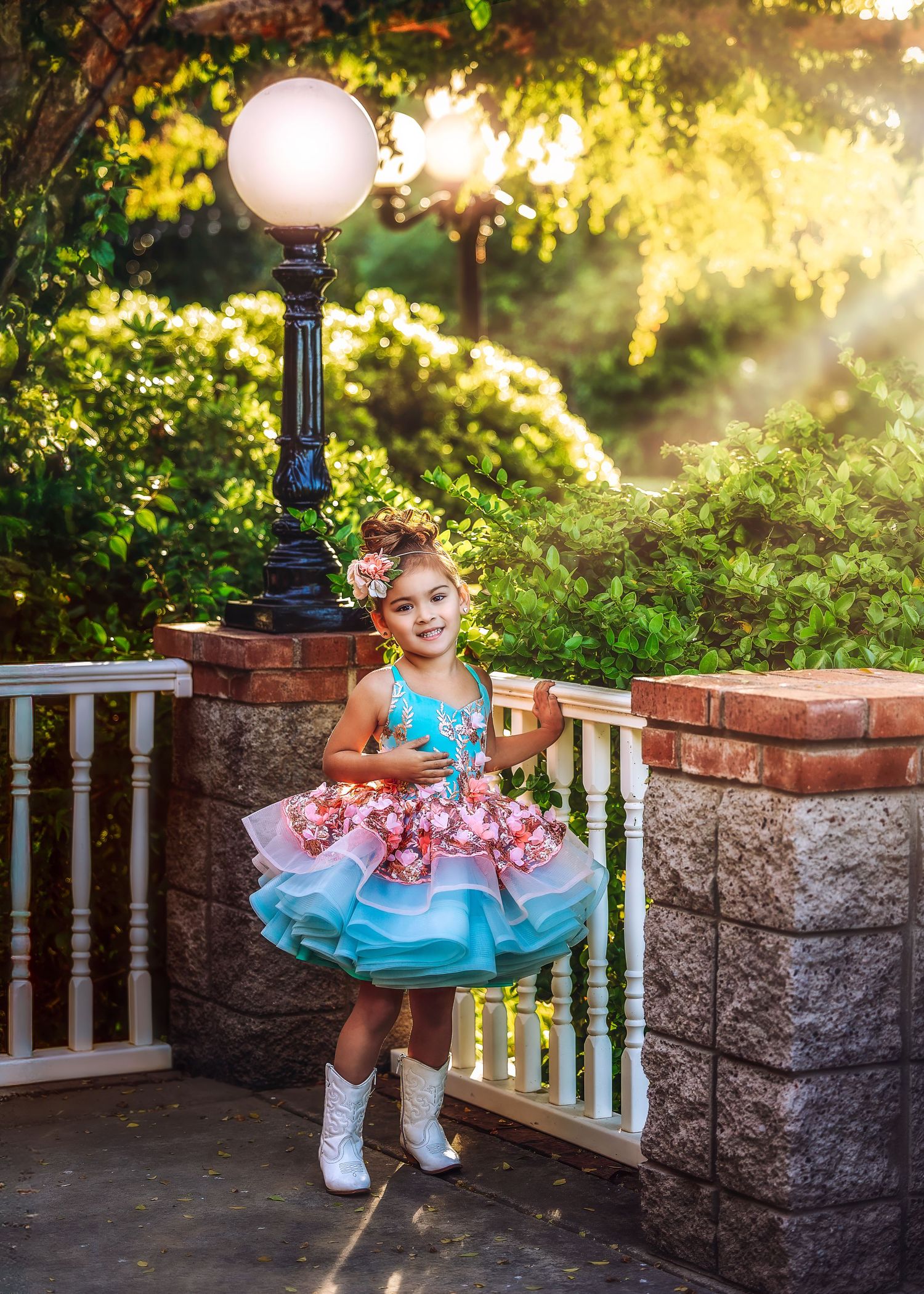 A little girl in a floral tiered dress and white boots poses by a glowing lamp post in a garden.