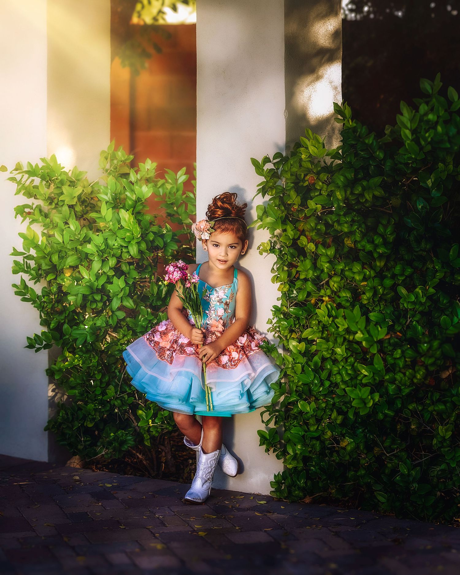 A little girl in a teal ruffled dress and white boots holds flowers while standing between lush green bushes.