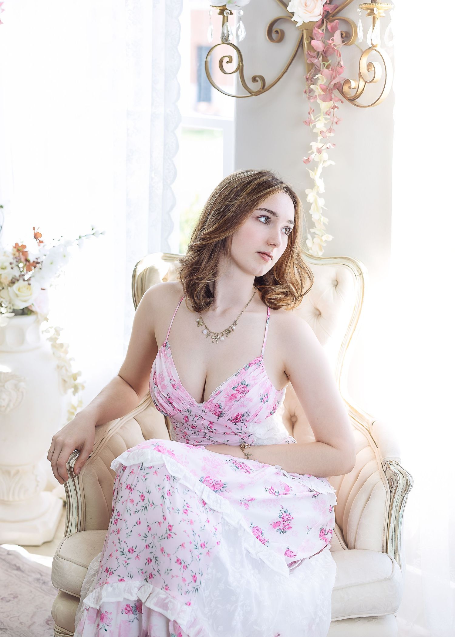 Brunette woman in pink floral dress with necklace seated in cream armchair by bright window.