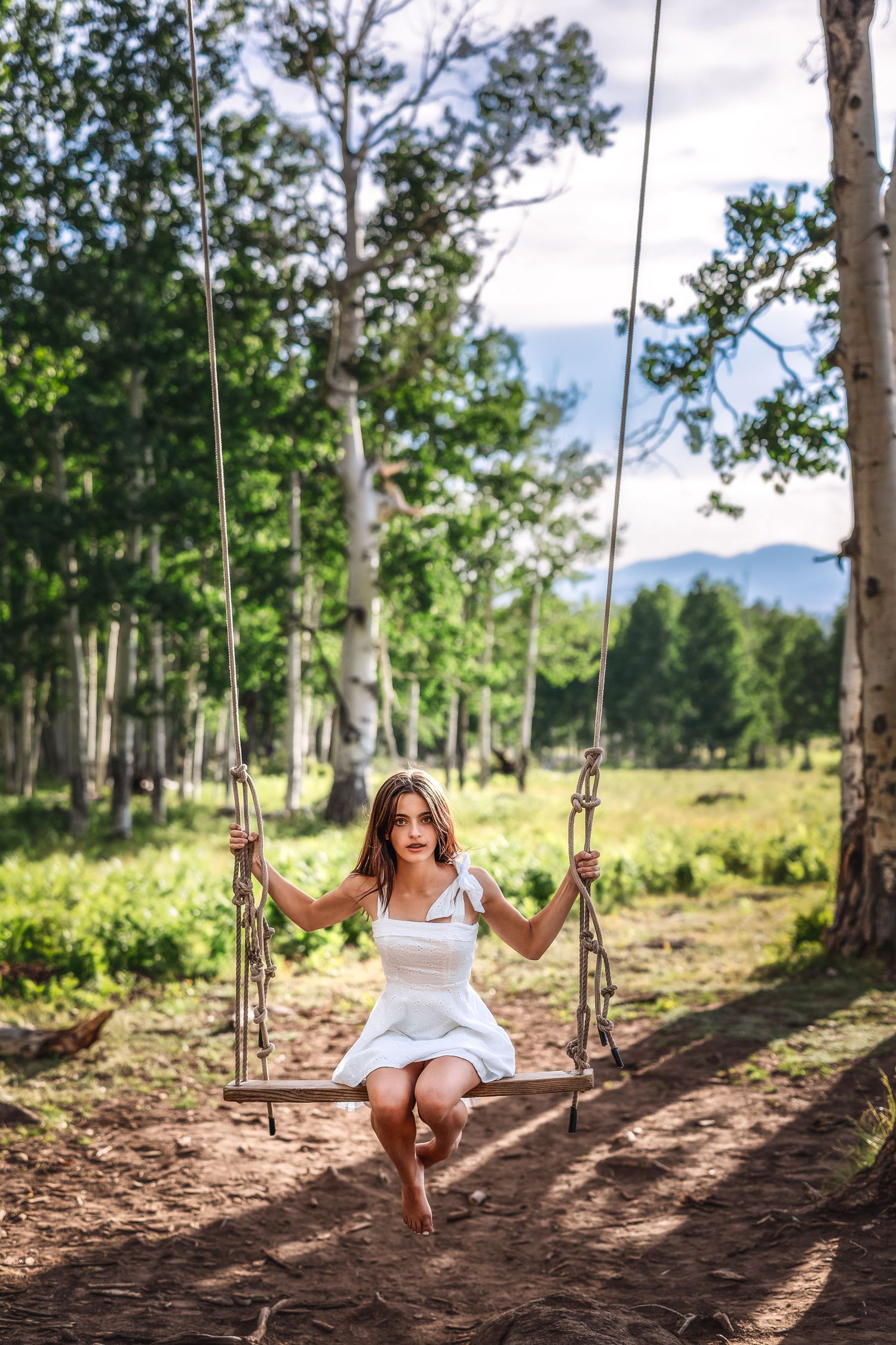 Young woman in white dress sitting on a rustic rope swing surrounded by lush green trees and mountains in background.