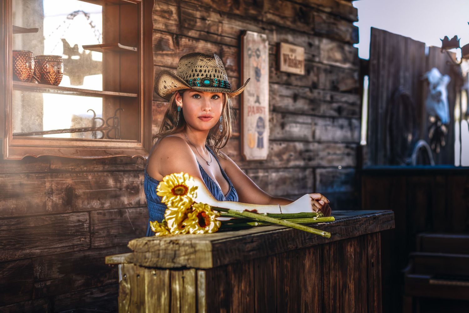 Young Asian woman in straw hat holding sunflowers at rustic wooden bar in western-style setting.