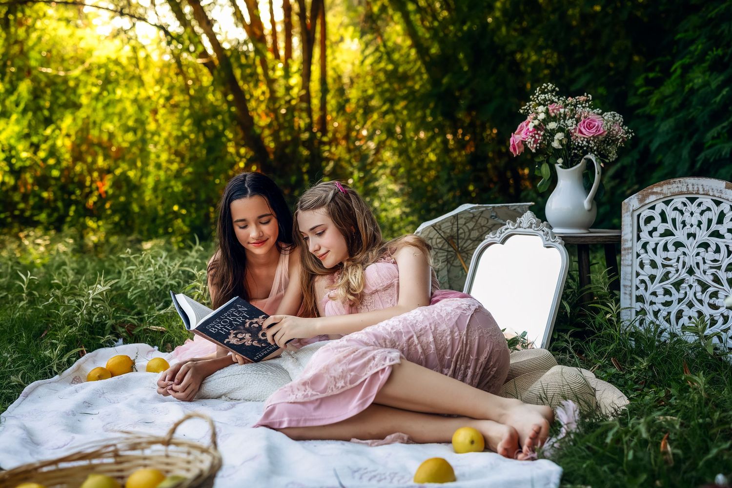 Two girls in pink dresses reading a book together on a blanket outdoors with lemons and flowers nearby.