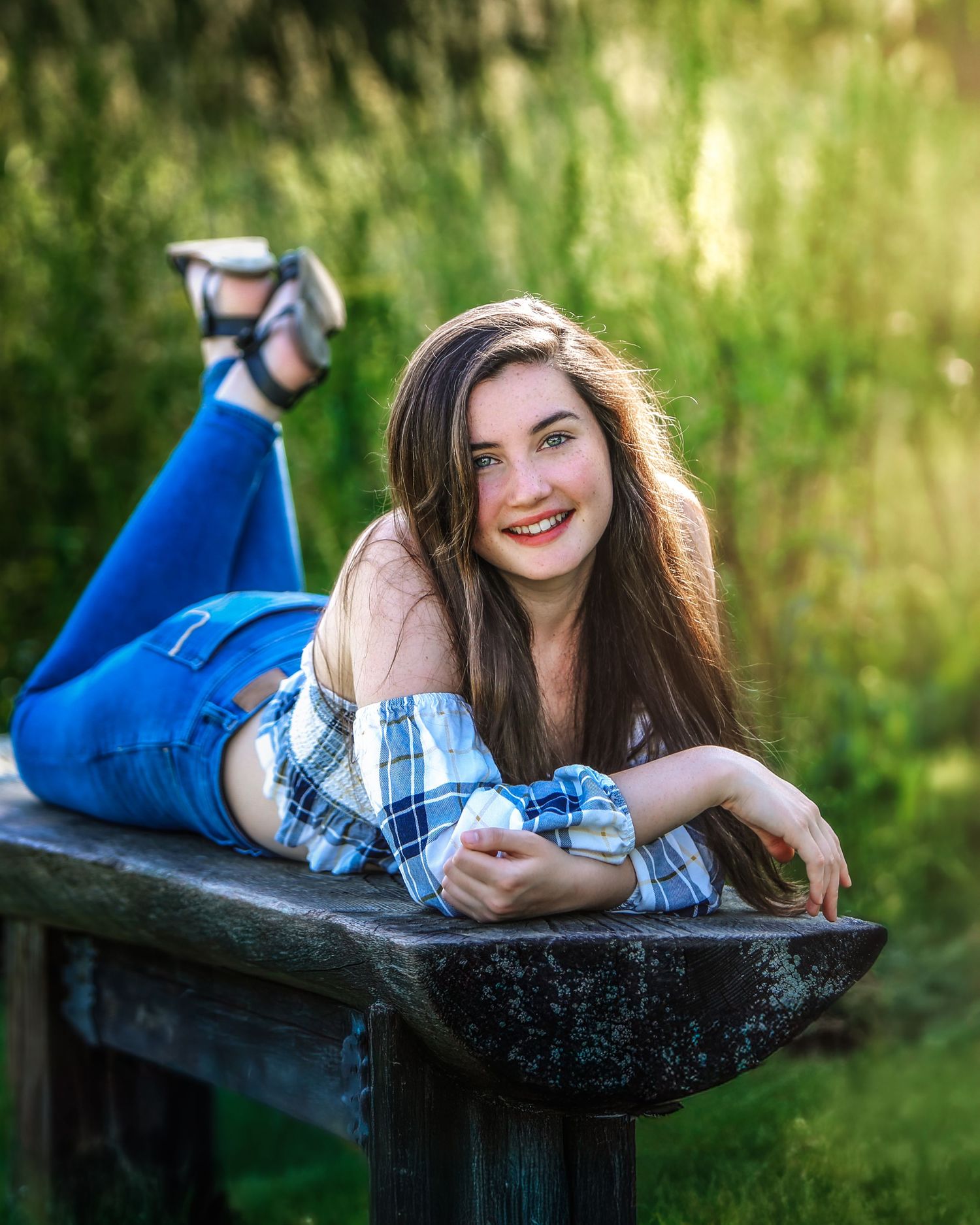 Smiling teenage girl in plaid shirt and jeans lying on rustic wooden bench outdoors.