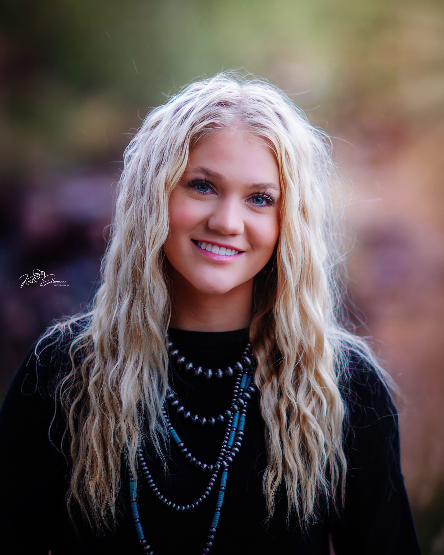 Portrait of smiling blonde teen girl with curly hair wearing black sweater and layered turquoise necklaces outdoors.