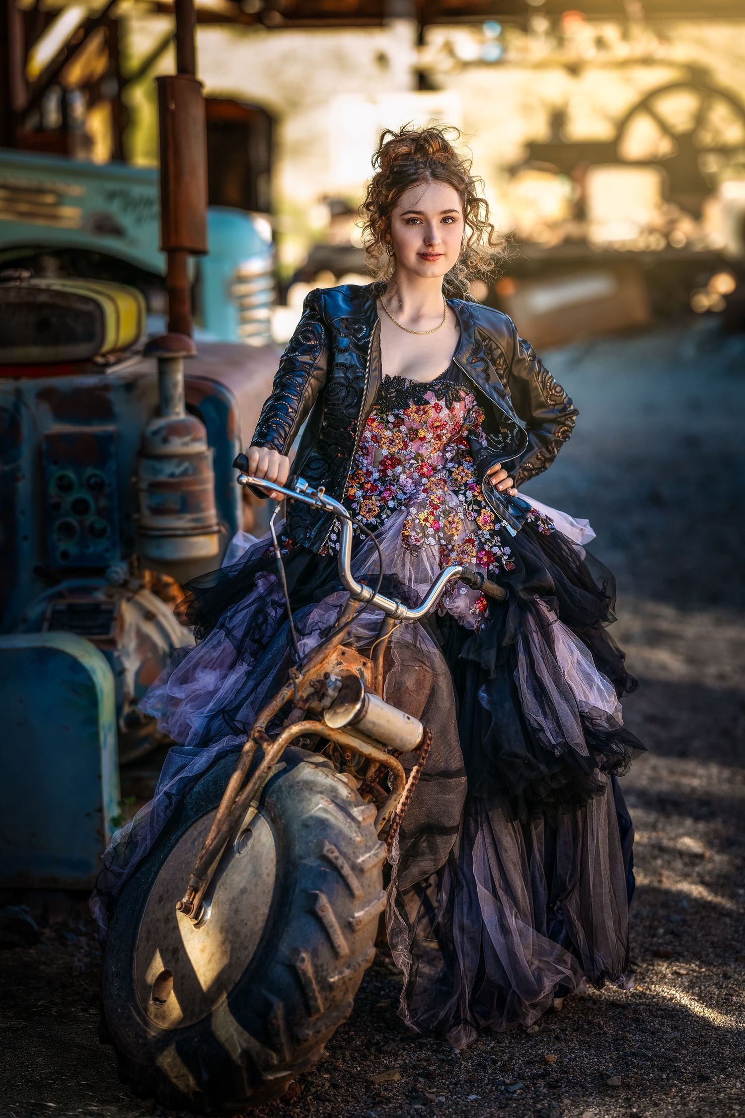 Young woman in floral corset, leather jacket, and purple tulle skirt poses with vintage motorcycle in rustic junkyard setting.