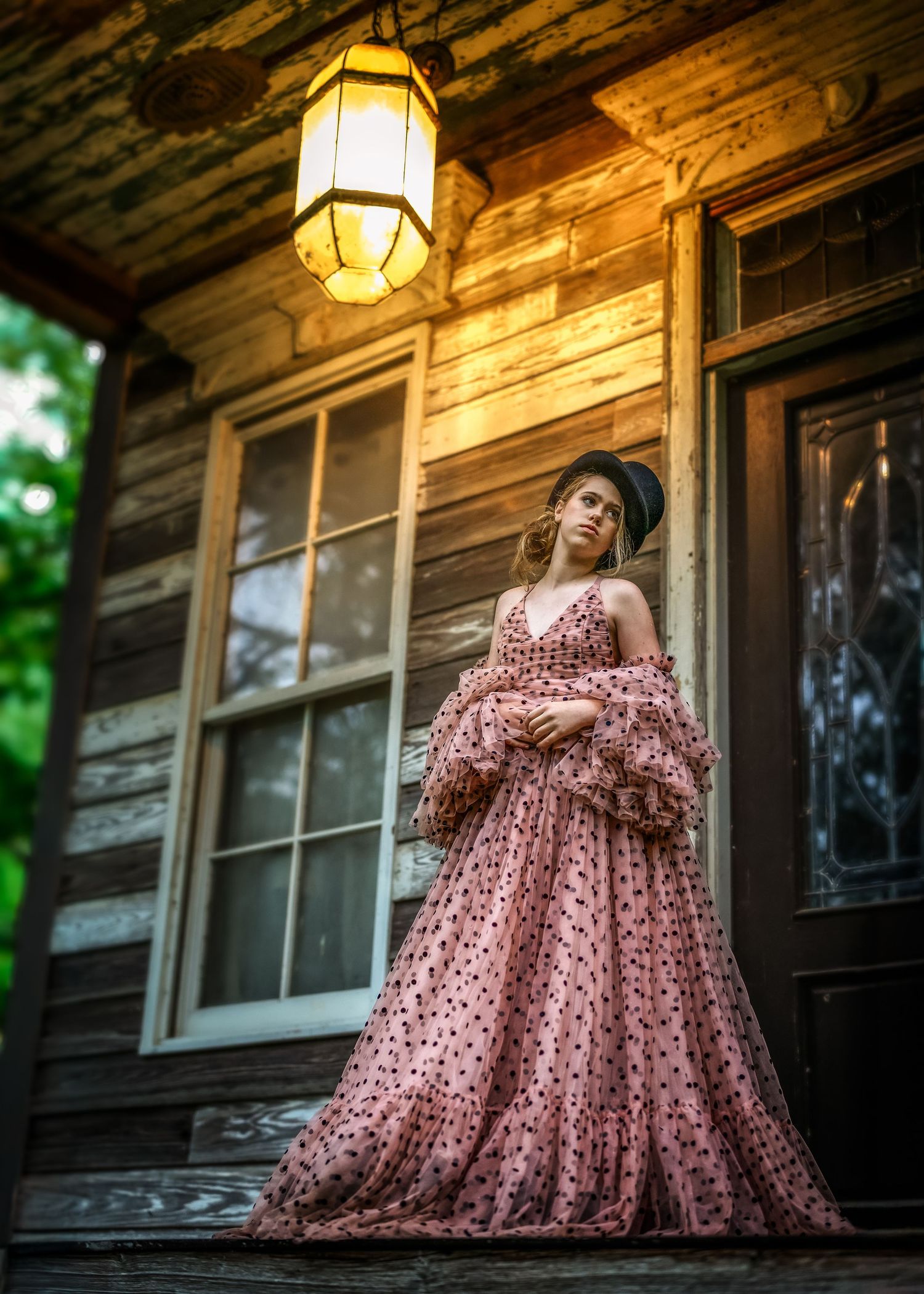 Woman in pink polka dot ball gown and black hat poses on rustic wooden porch under glowing lantern.