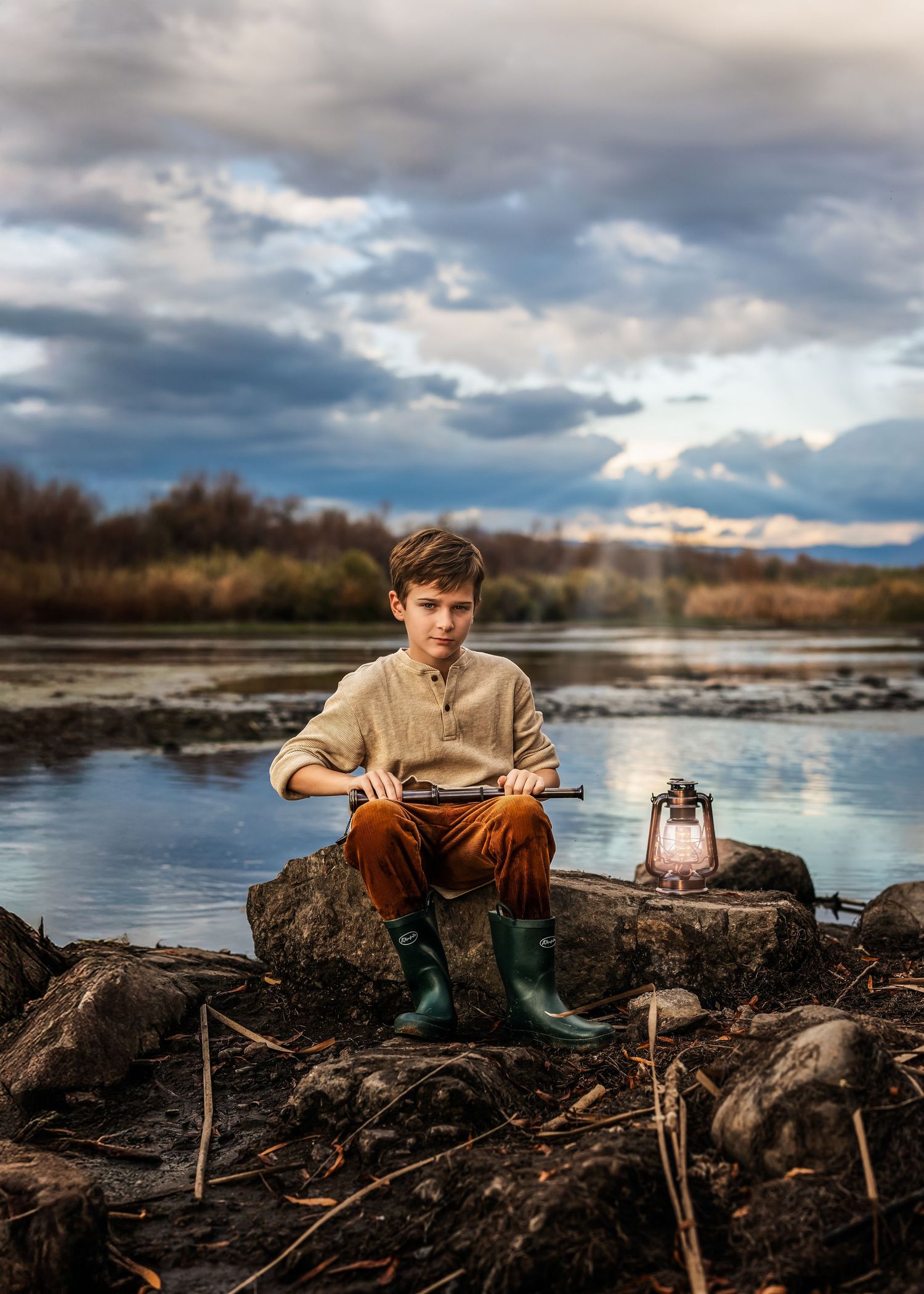 Boy in linen shirt and green boots sits on rocks by a lake holding a telescope with a lantern nearby.