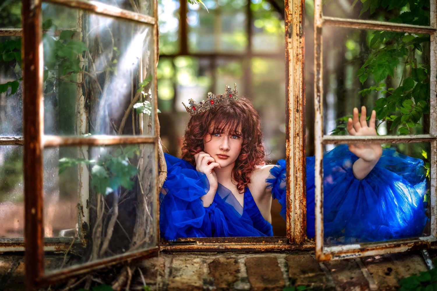 Woman in blue gown poses in rustic broken greenhouse window surrounded by lush green foliage.