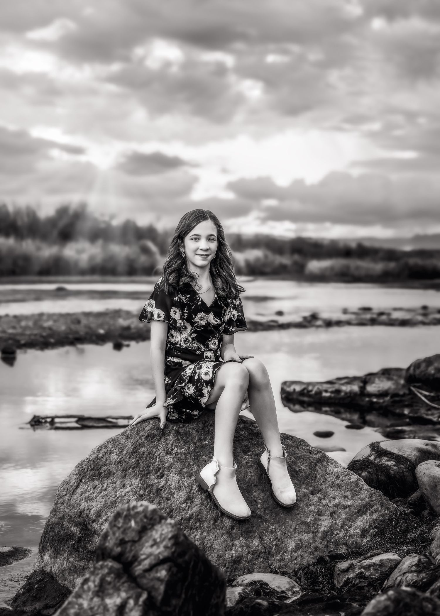 Black and white portrait of a teenage girl sitting on a rock by a river at sunset.