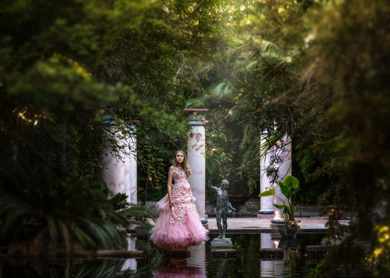 Elegant young woman in pink ball gown stands in a lush garden with classical columns and statues.