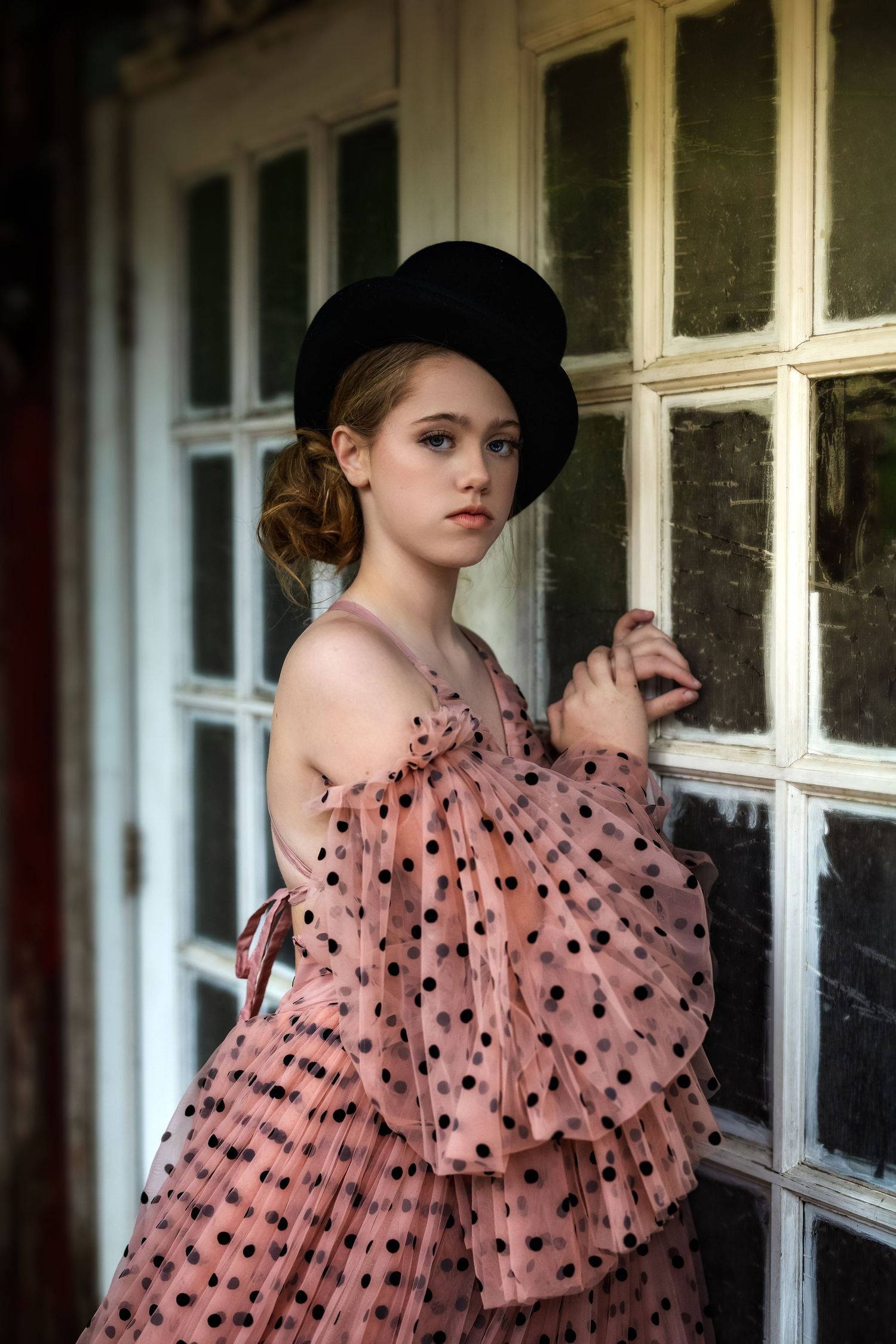 Young woman in pink polka dot ruffled dress and black top hat posing elegantly by vintage white-framed windows.