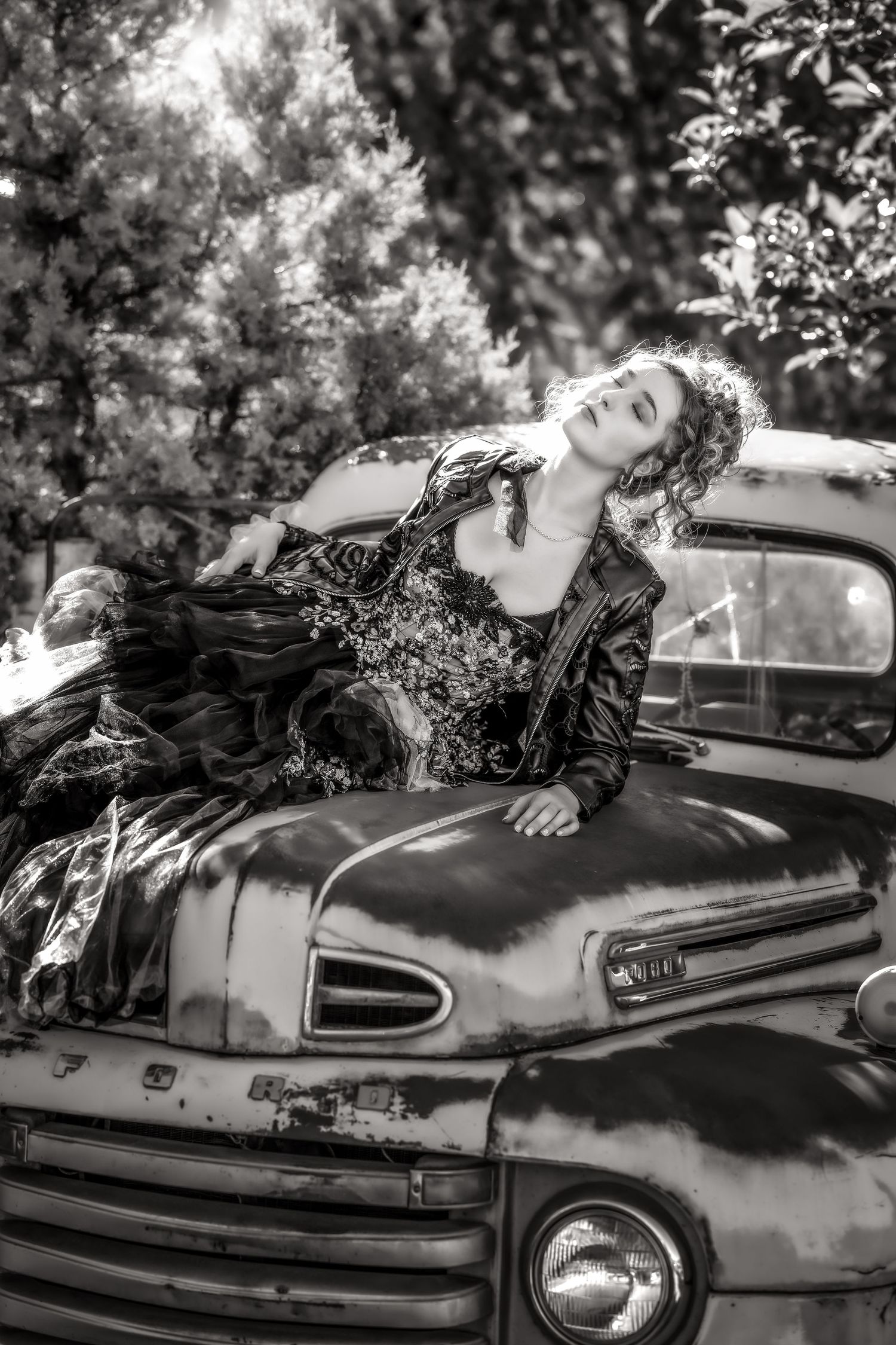 Black and white photo of a woman in a vintage gown reclining on an old rustic truck hood surrounded by lush trees.