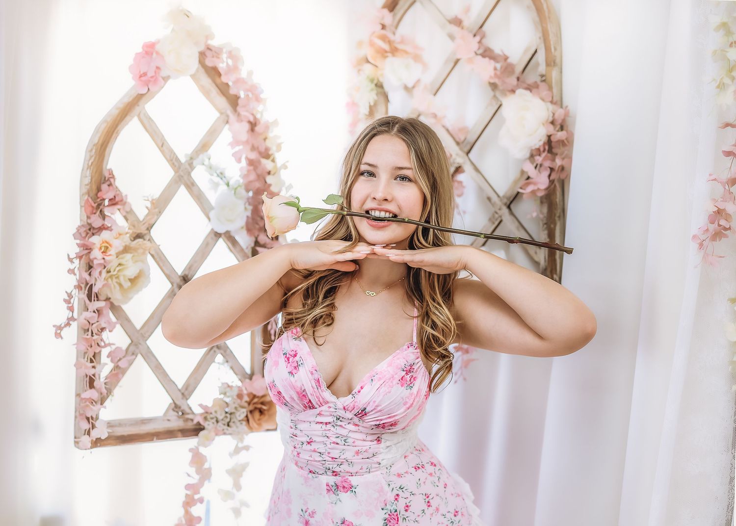 Playful young woman in pink floral dress posing with a rose stem held in her mouth outdoors.