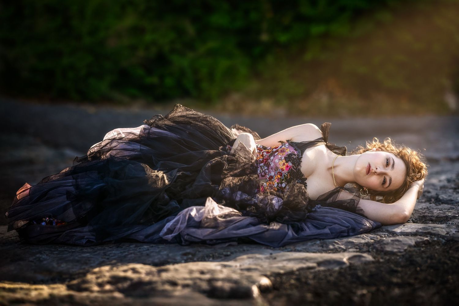 A woman in a dark floral gown lies gracefully on rocky ground in a dreamy, ethereal outdoor setting.