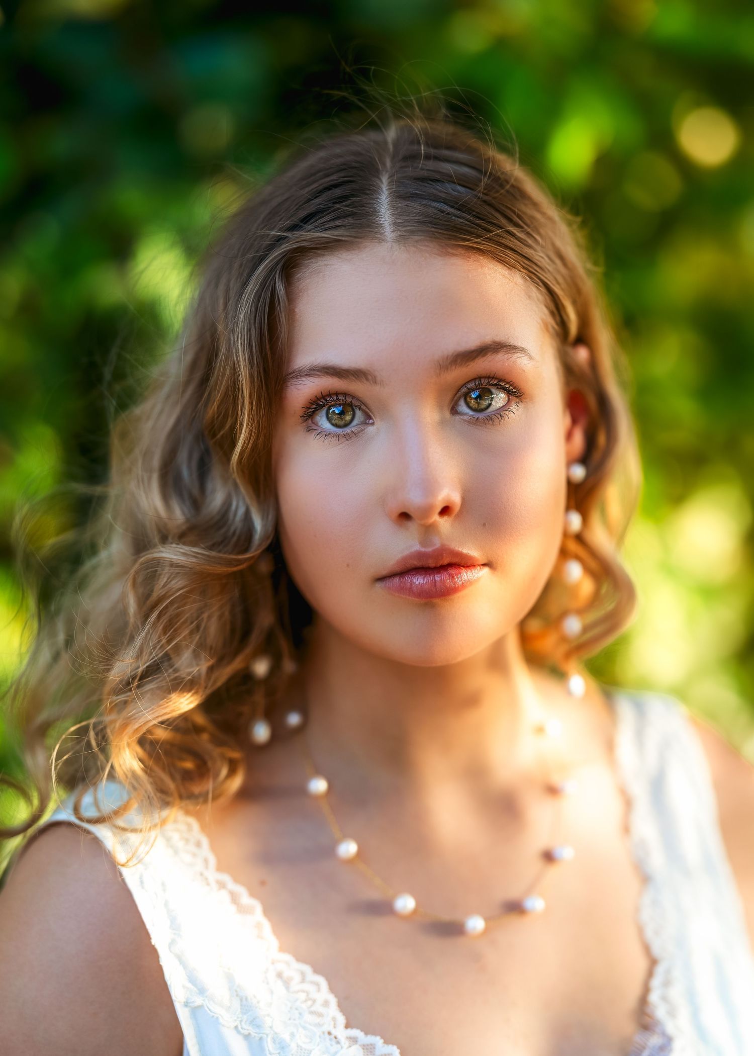 Young woman with wavy hair and green eyes wearing a white dress and pearl necklace, posing outdoors with bokeh background.