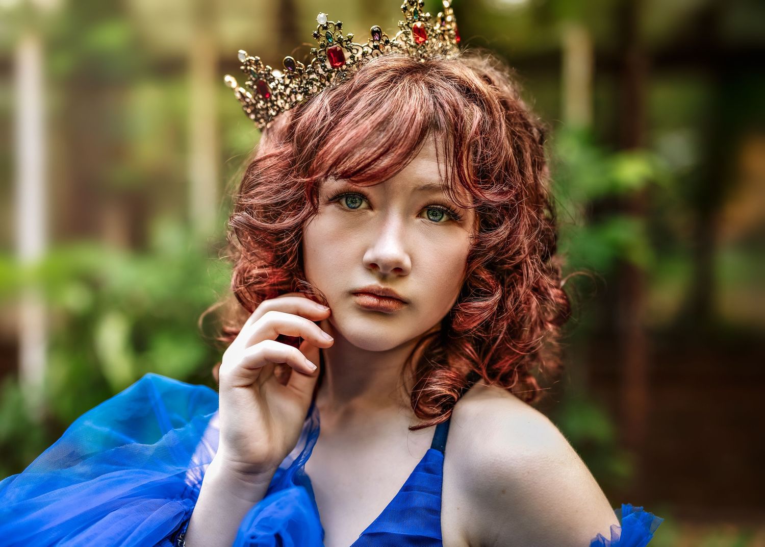 Young woman with red curly hair wearing a gold crown and blue dress poses gracefully outdoors.