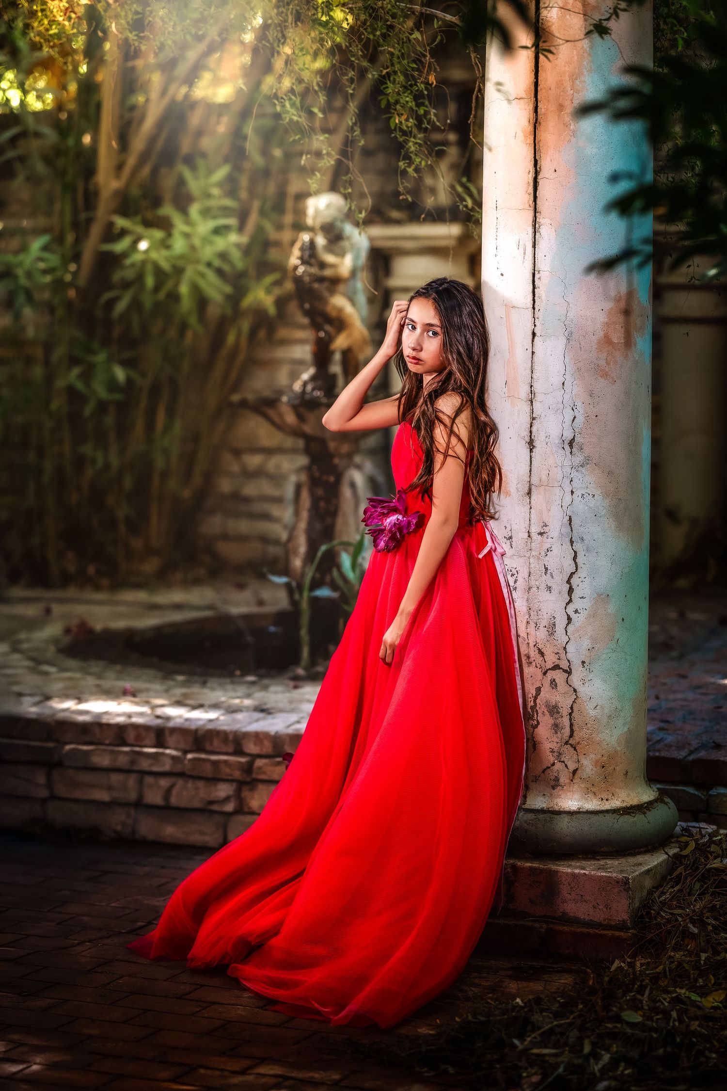 Girl in flowing red ball gown stands by weathered column in lush garden with bronze statue.