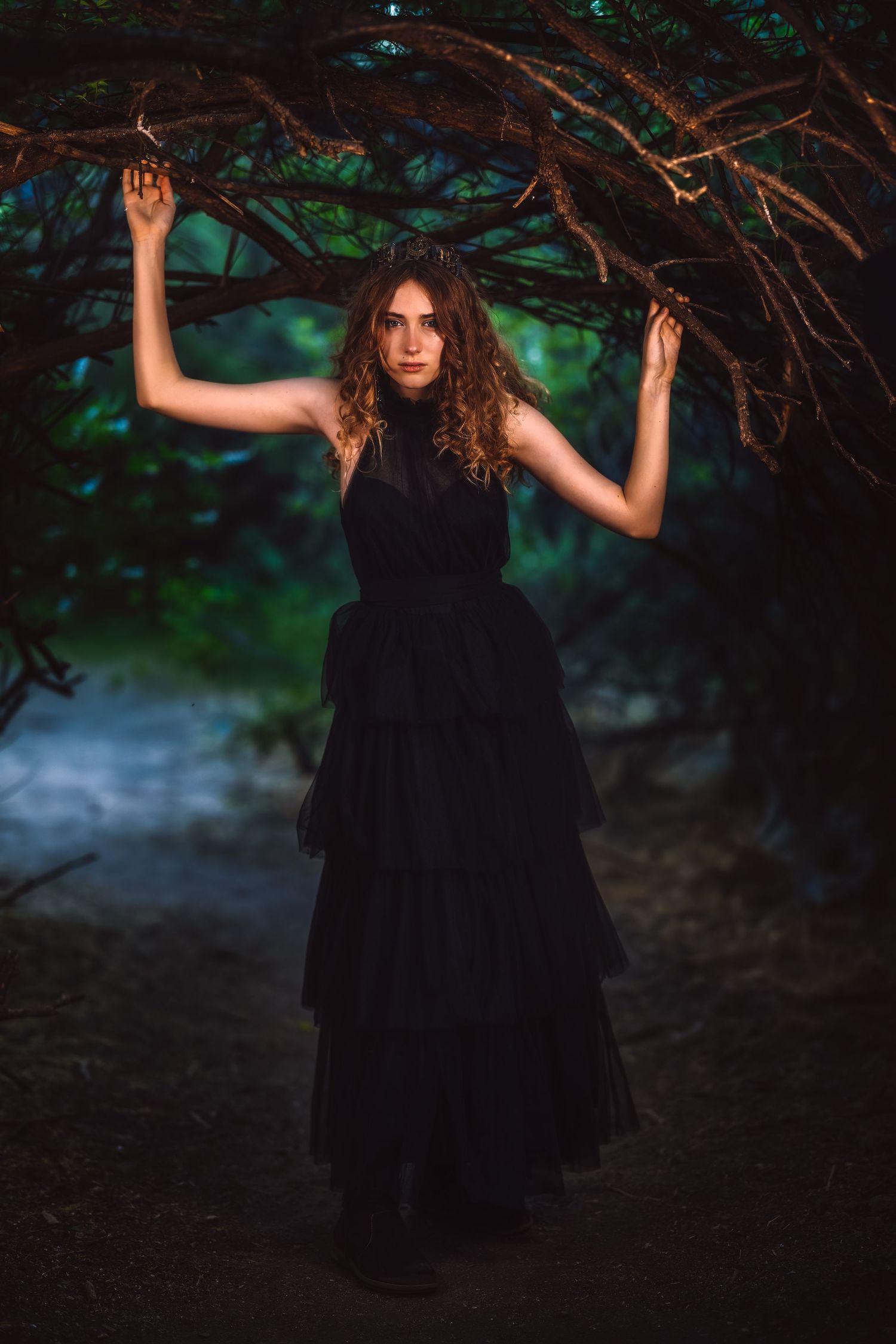 Woman in black tiered gown poses dramatically under arching tree branches in a dark, moody forest setting.
