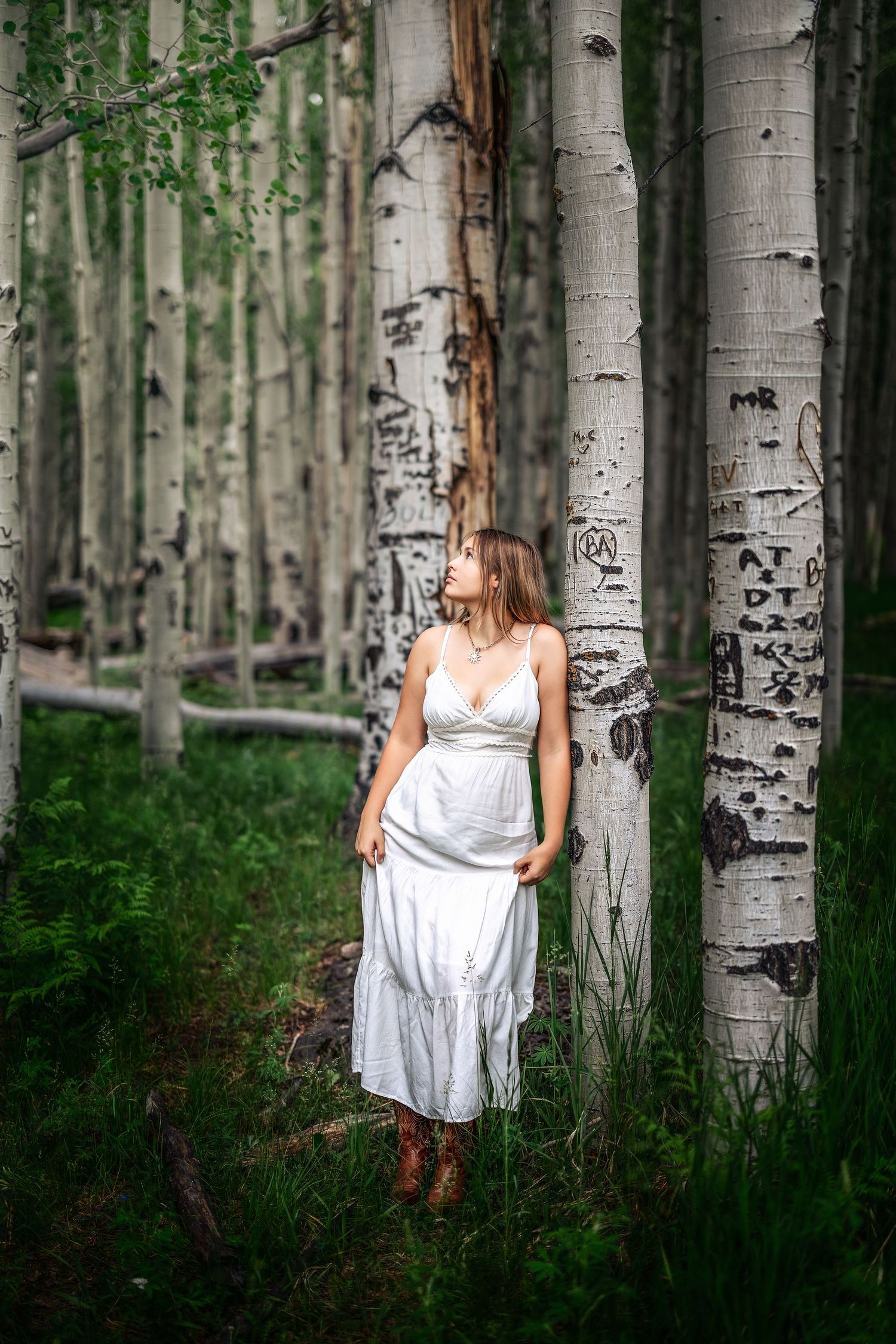 Woman in white dress and cowboy boots standing among aspen trees with carved bark in a lush green forest.