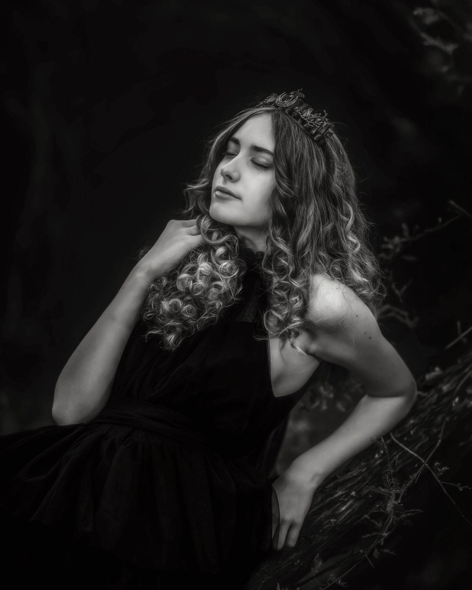 Black and white portrait of a young woman in a dark dress wearing a crown, posing gracefully outdoors.
