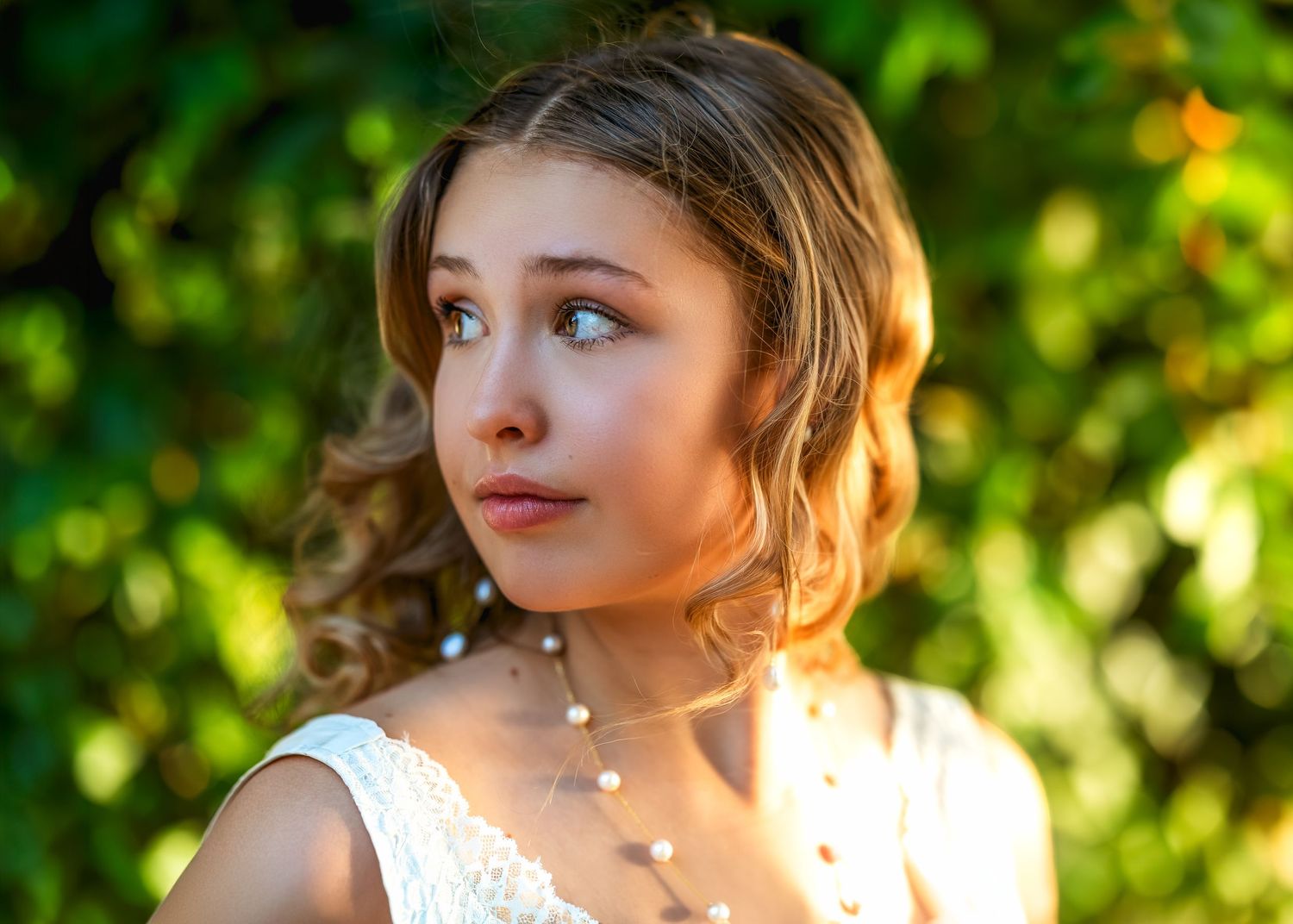 Young woman in white lace dress wearing pearl necklace, posing outdoors with soft sunlight and green foliage background.