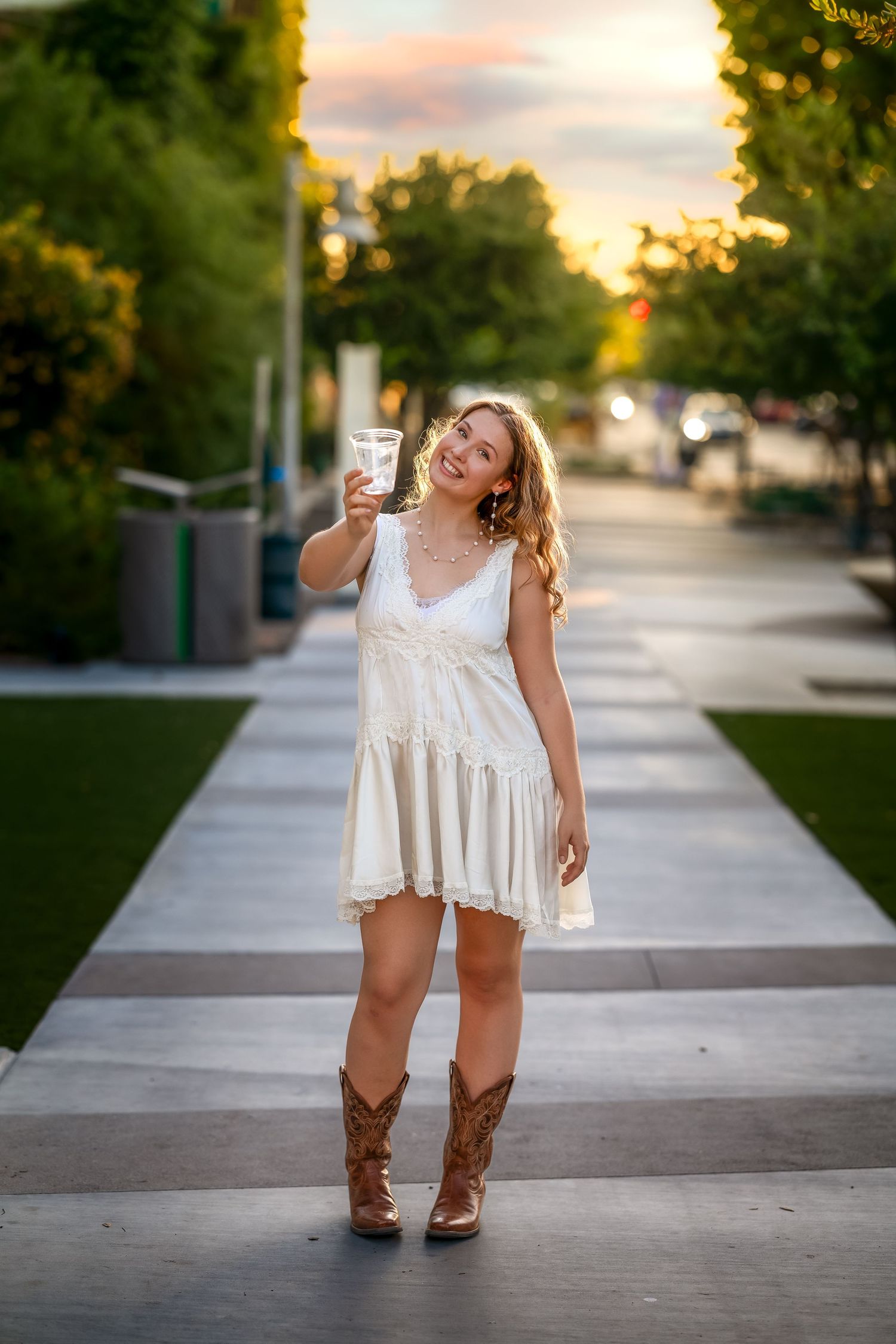 Young woman in white dress and cowboy boots holding a drink, smiling outdoors at sunset.