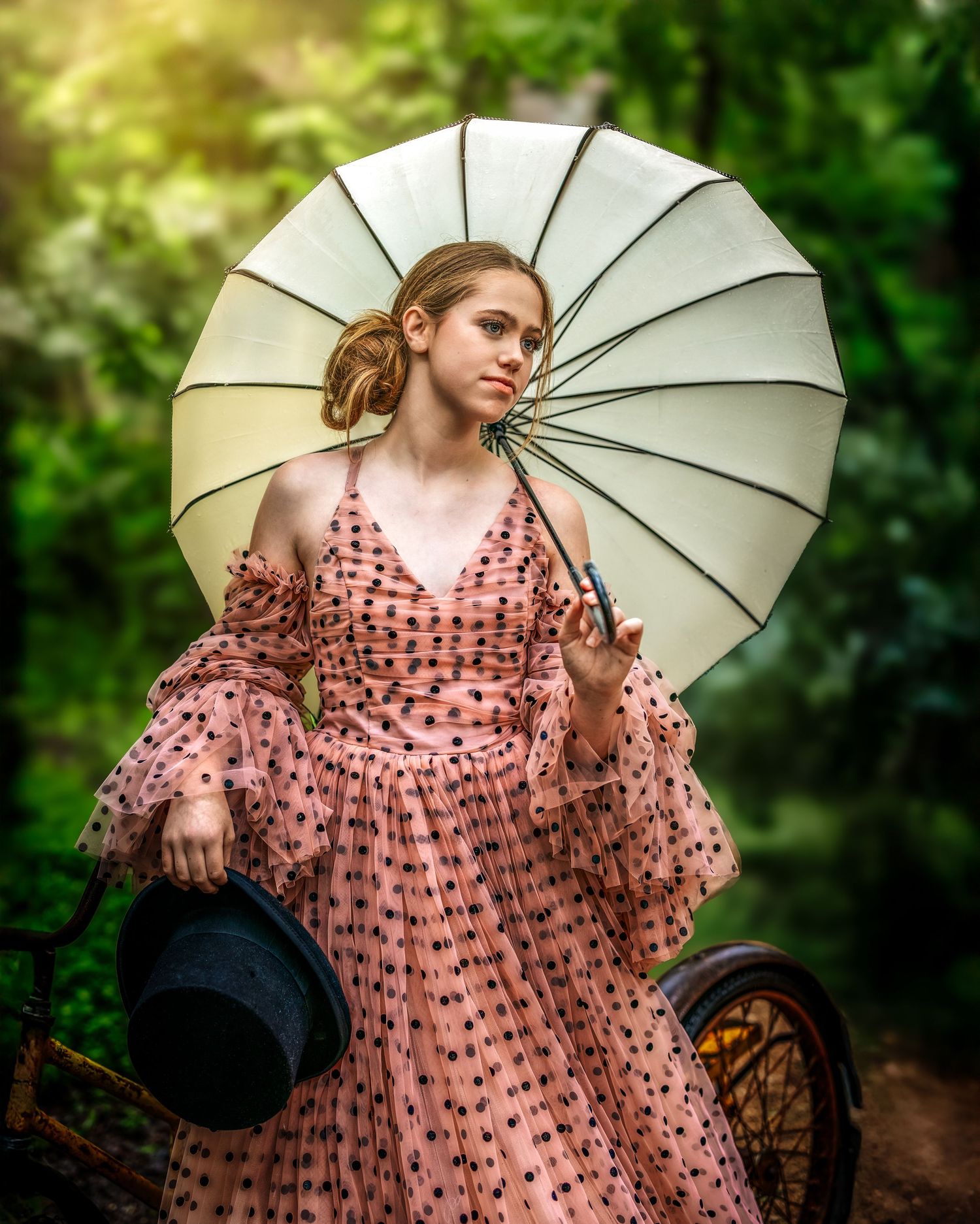 Young woman in pink polka dot dress holding parasol and top hat near bicycle in lush green forest setting.