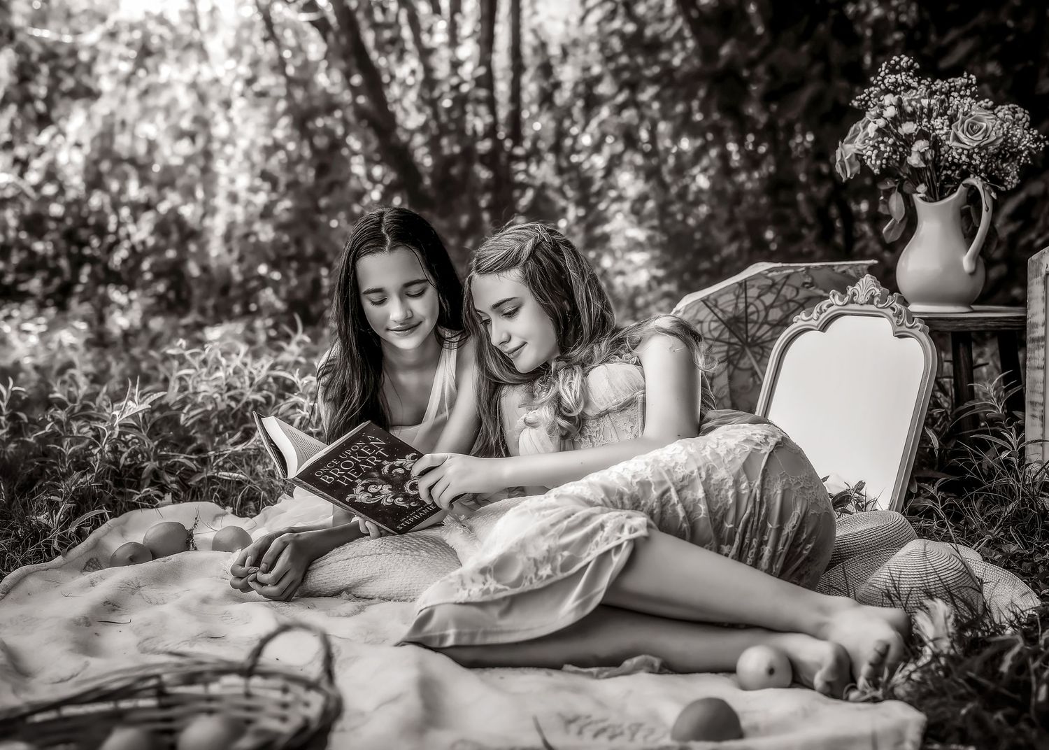 Two girls sitting outdoors on a blanket reading a book together in a vintage black and white photo.