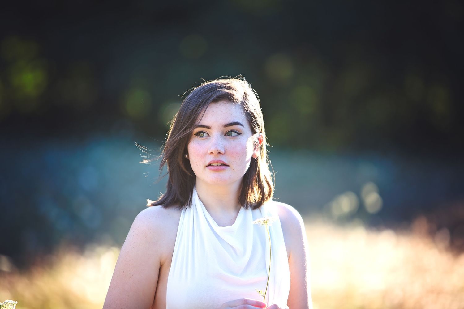 Young woman with short brown hair in white dress gazing upward outdoors in golden sunlight.