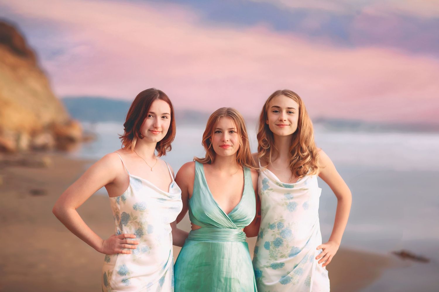 Three women in white and teal dresses smiling together on a beach at sunset.