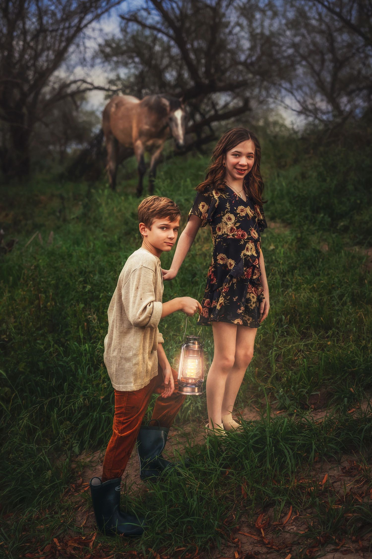 Two children hold a glowing lantern in a forest meadow with a horse grazing in the background at dusk.
